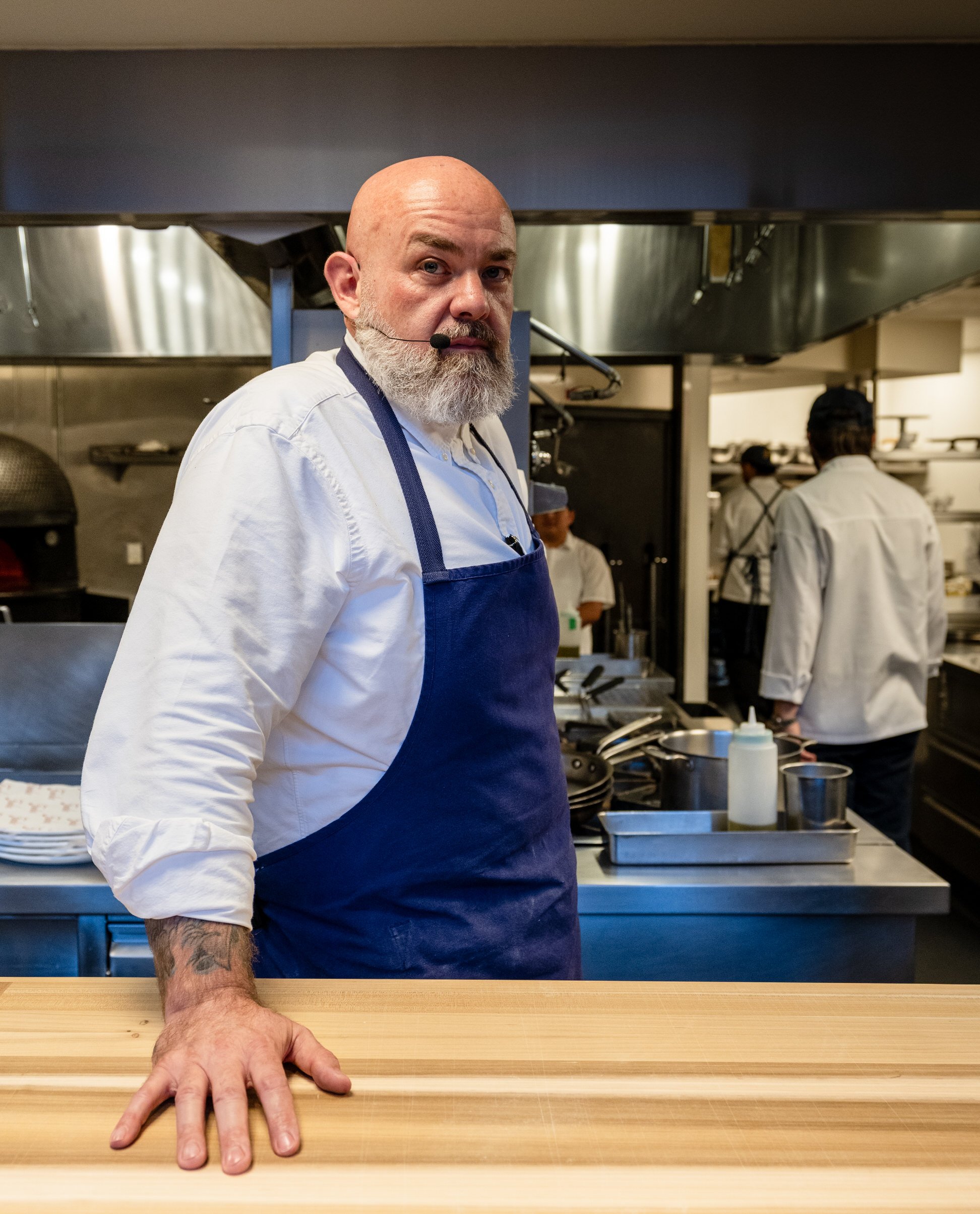 Chef Evan Fluke with a bald head, gray beard, white shirt, blue apron, and headset microphone stands behind a wooden counter in a commercial kitchen, with other chefs working in the background at FX's The Bear event.