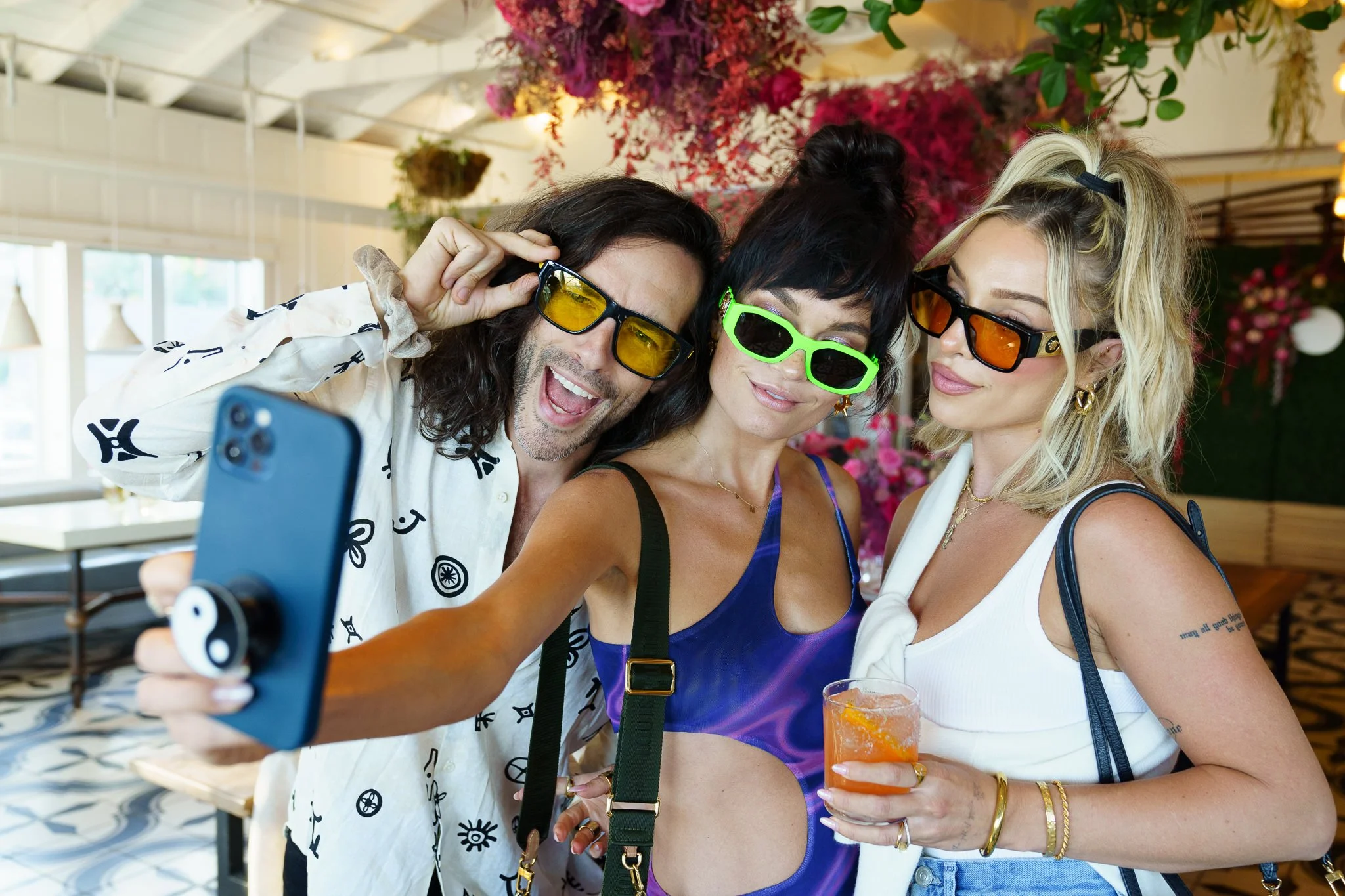 Three friends taking a selfie together at a social gathering, wearing colorful sunglasses and smiling, with a floral background and a drink in one person's hand at Sports Illustrated Swimsuit Issue event in Miami.