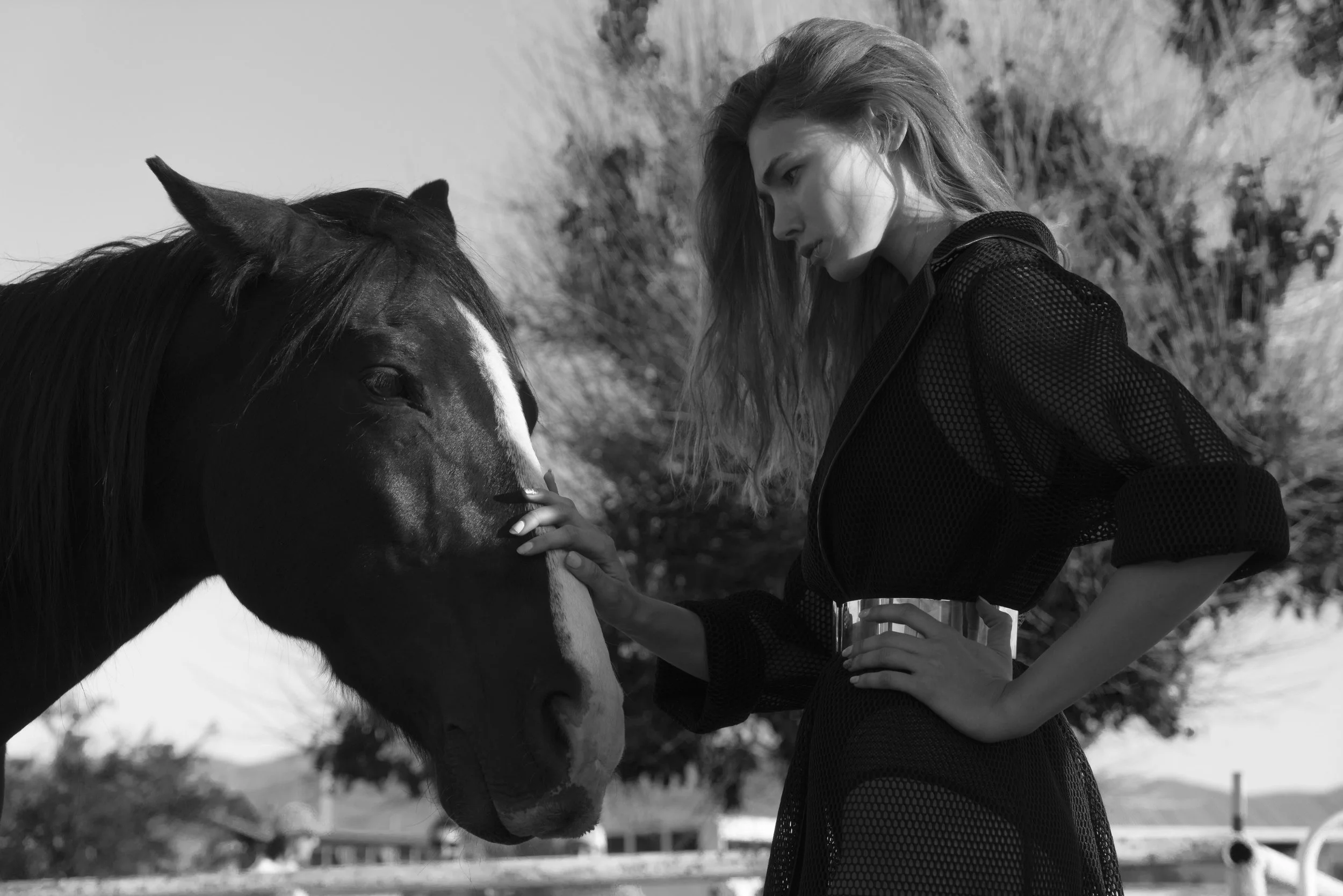 A woman with long hair in a mesh top feeding a horse in an outdoor setting.