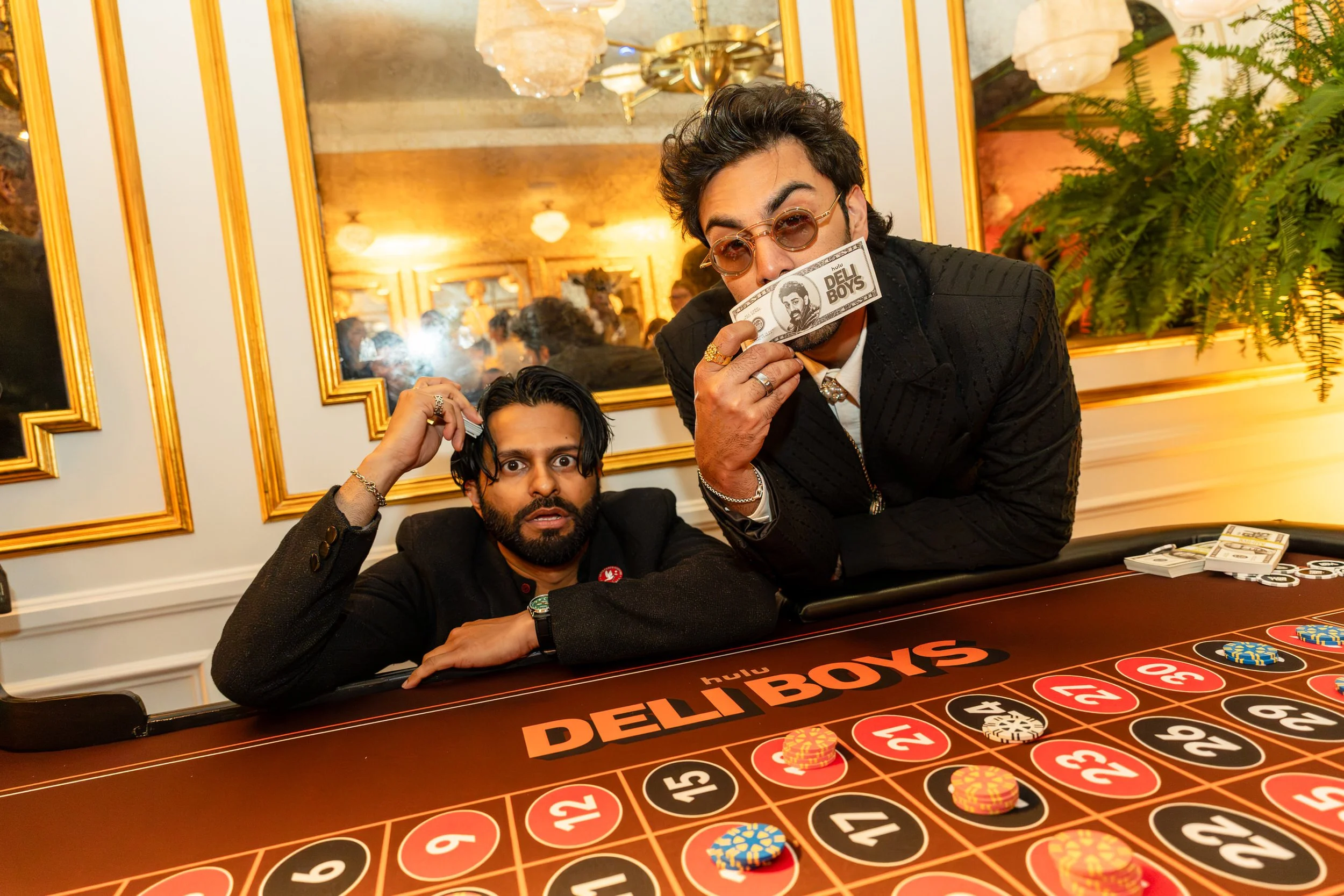 Two men are sitting at a casino table with a roulette wheel. One man is holding chips and looking surprised, while the other is holding a stack of money with the words 'DEL BOYS' printed on it, and wearing sunglasses. The background features a luxuri
