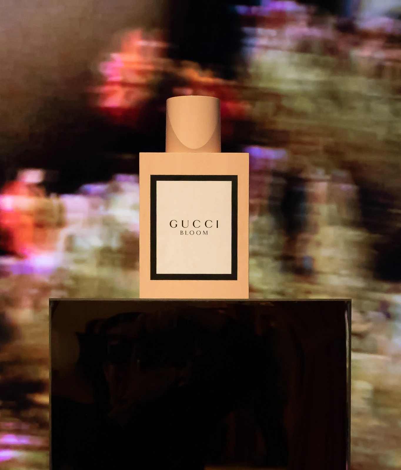 A bottle of Gucci Bloom perfume on a stand with a dark, reflective surface, with a blurred colorful background.
