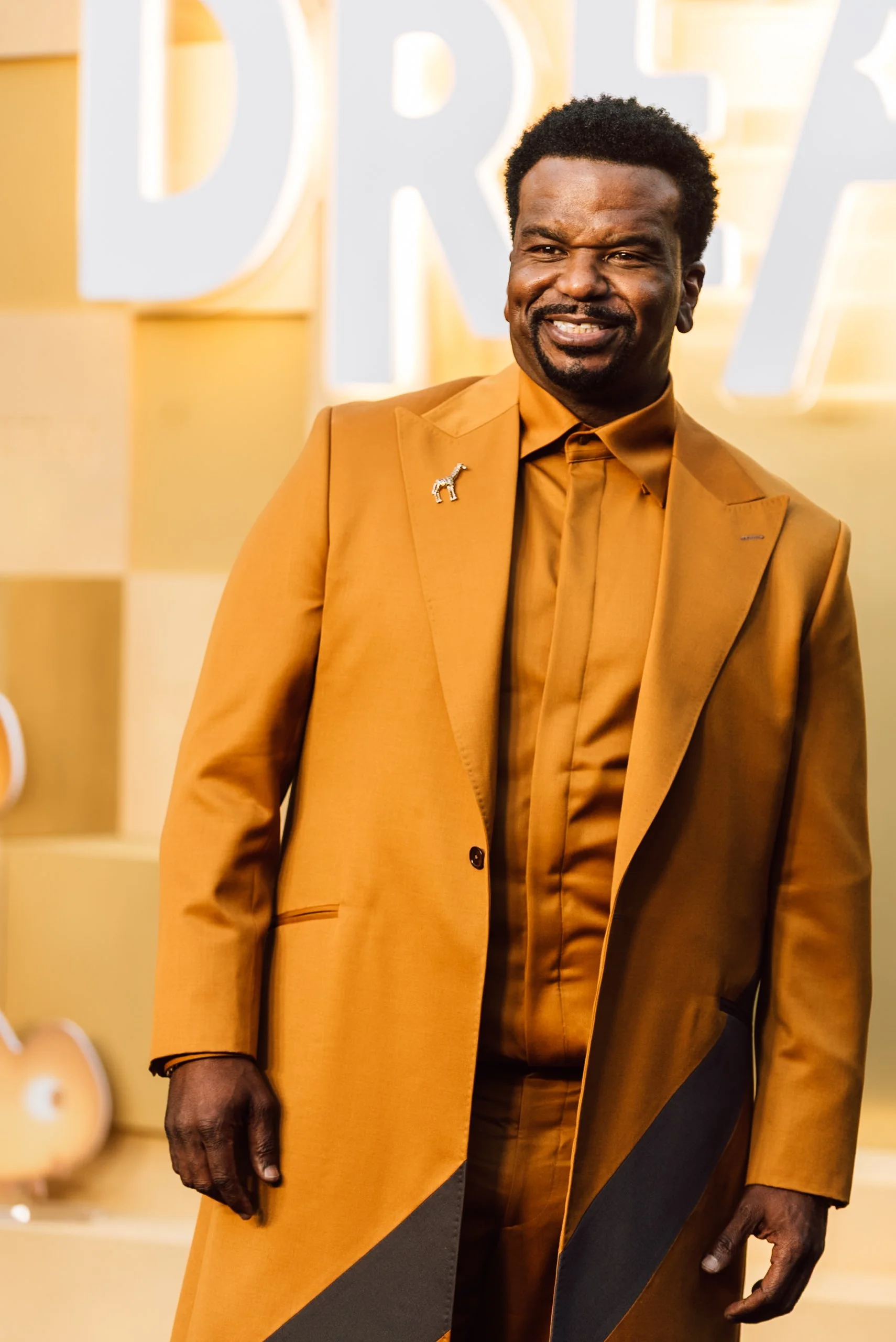 Craig Robinson smiling, wearing a mustard blazer with a giraffe-shaped brooch, mustard shirt, and dark pants, standing in front of a golden background for NETFLIX's "In Your Dreams" movie premiere.