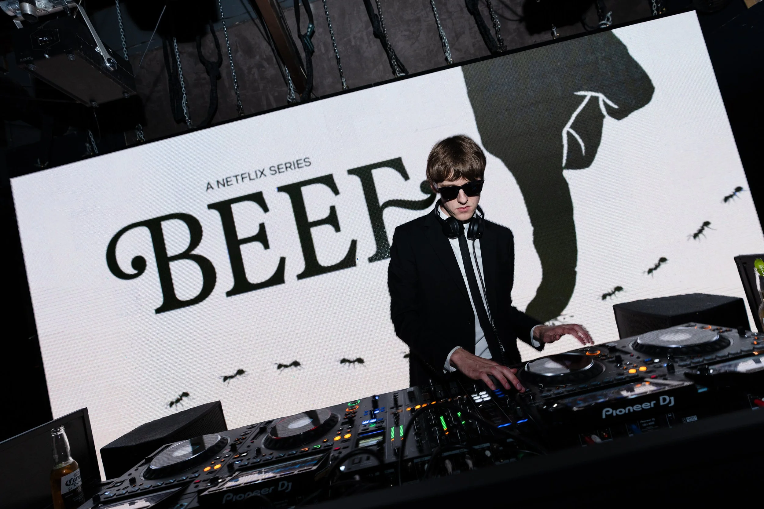 Producer and DJ The Dare wearing sunglasses and headphones performing at a nightclub, with a large screen displaying the title of a Netflix series and small images of ants in the background at NETFLIX's Beef Season 2 premiere party.