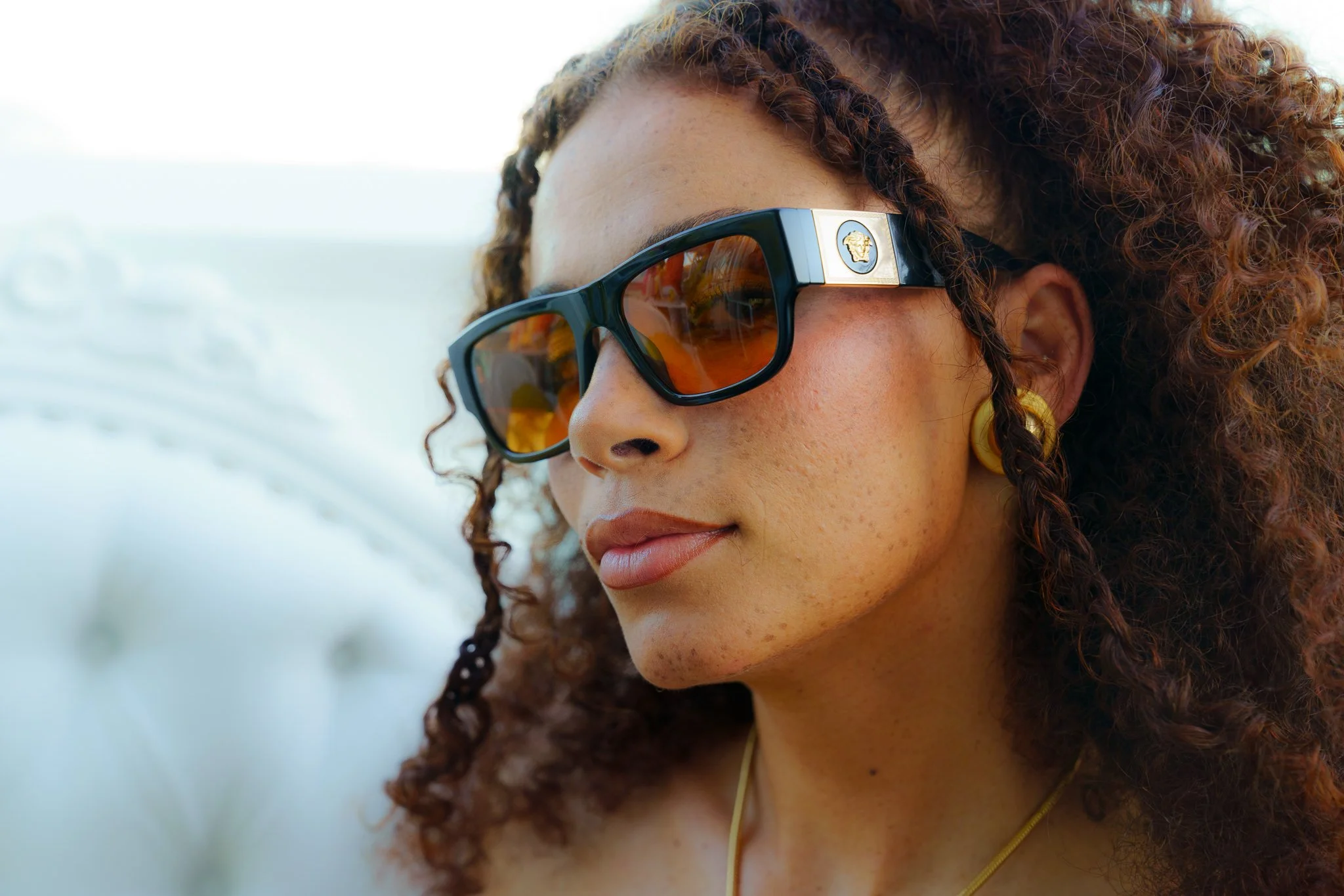 Close-up of a woman with curly hair wearing sunglasses with a gold emblem and a yellow earring, outdoors with a blurry sky background. Sunglass Hut.