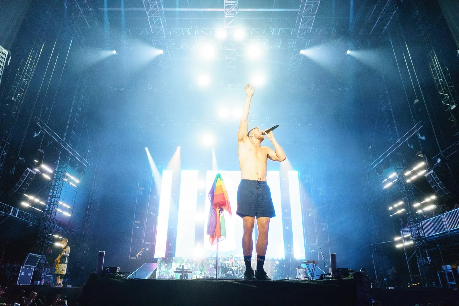 Dan Reynolds from Imagine Dragons holding a microphone and raising his right hand on a brightly lit stage with LED screens and multicolored rainbow flags in front of him.