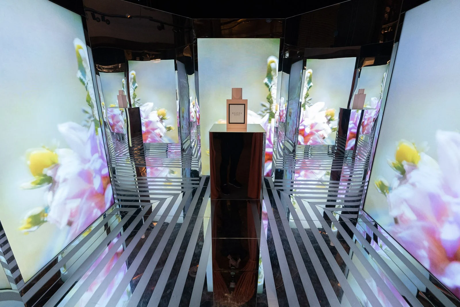 An illuminated display with multiple screens showing pink and yellow flowers, a centered Gucci perfume bottle on a glossy table, and a mirrored reflective floor and walls creating a tunnel effect.