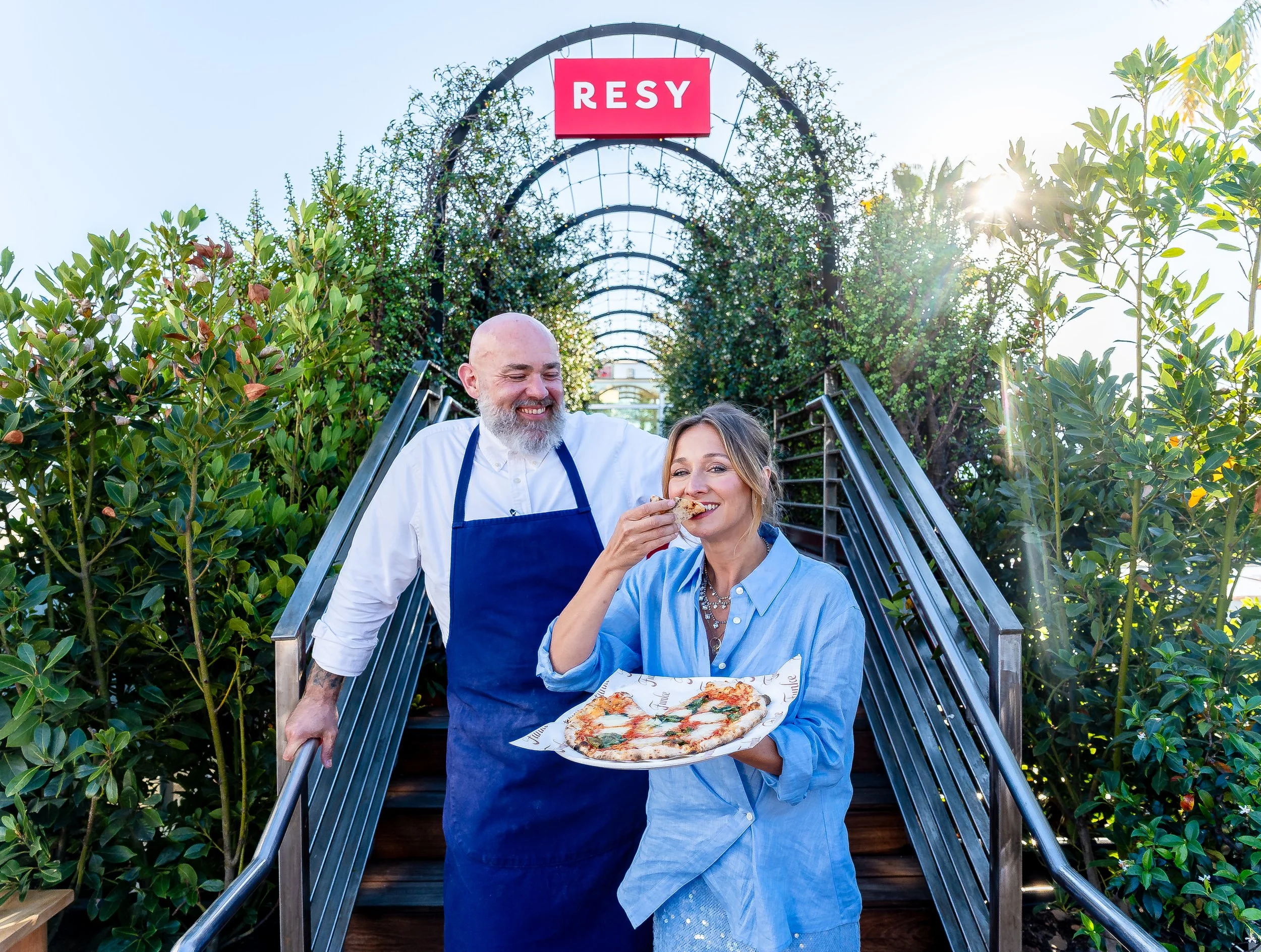Chef Evan Fluke  wearing a white chef's coat and blue apron, helping a woman who is eating a slice of pizza. They are on an outdoor staircase surrounded by green foliage with sunlight shining through at FX's The Bear event.