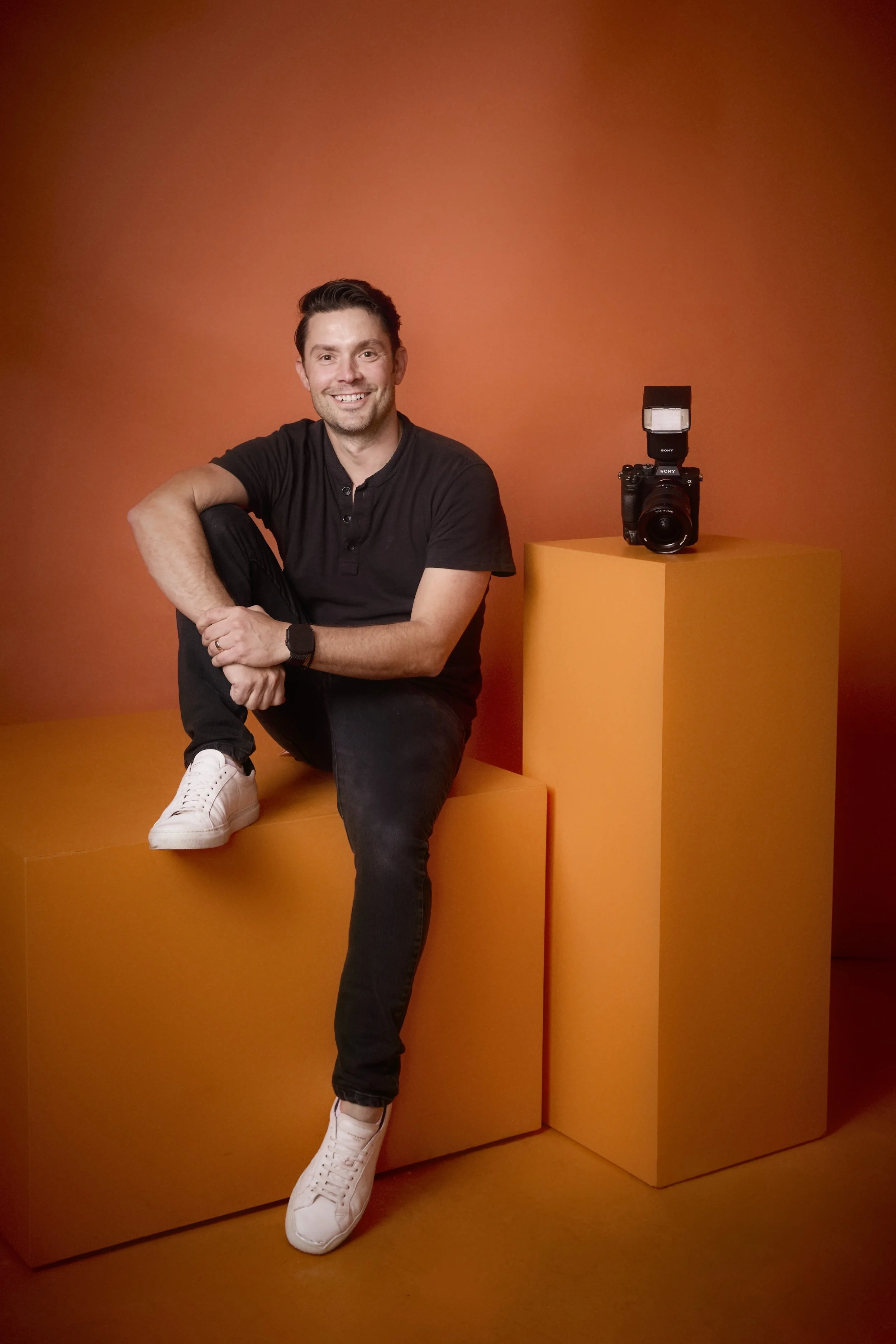 A man with short dark hair, wearing a black shirt and black pants, sitting on orange blocks against an orange background, smiling at the camera. A camera with a flash is placed on one of the orange blocks beside him.