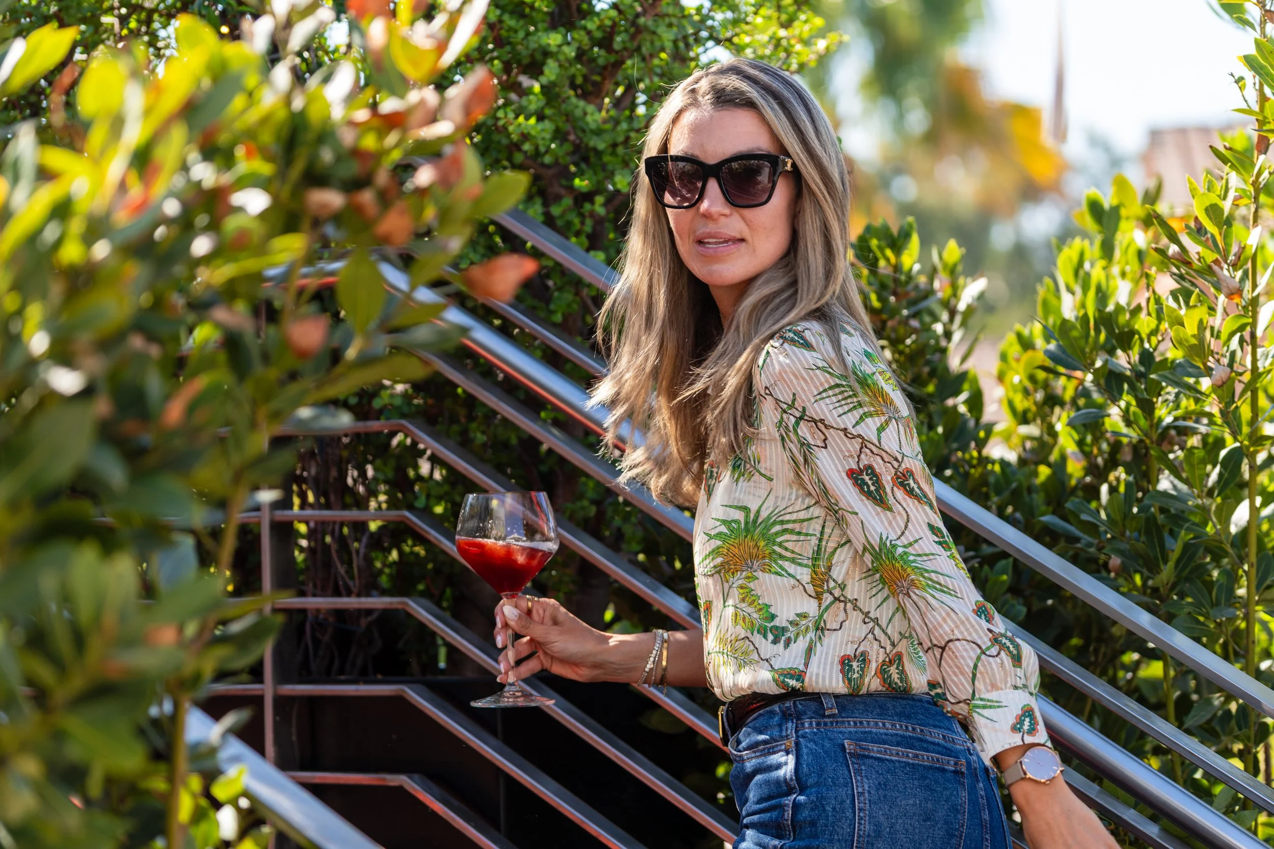 A woman with blonde hair wearing sunglasses and a tropical-patterned shirt holding a glass of red wine outdoors with green bushes in the background.