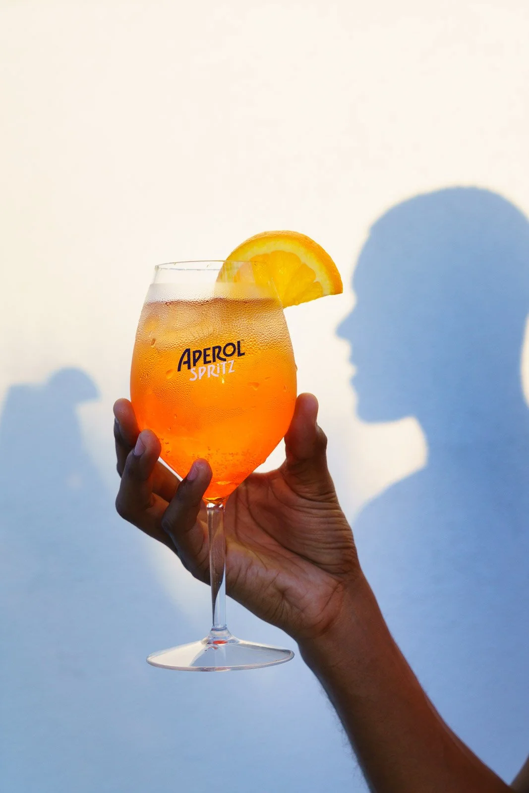 A hand holding a glass of orange Aperol Spritz with a lemon wedge garnish, casting a shadow of a woman's profile on a white wall.