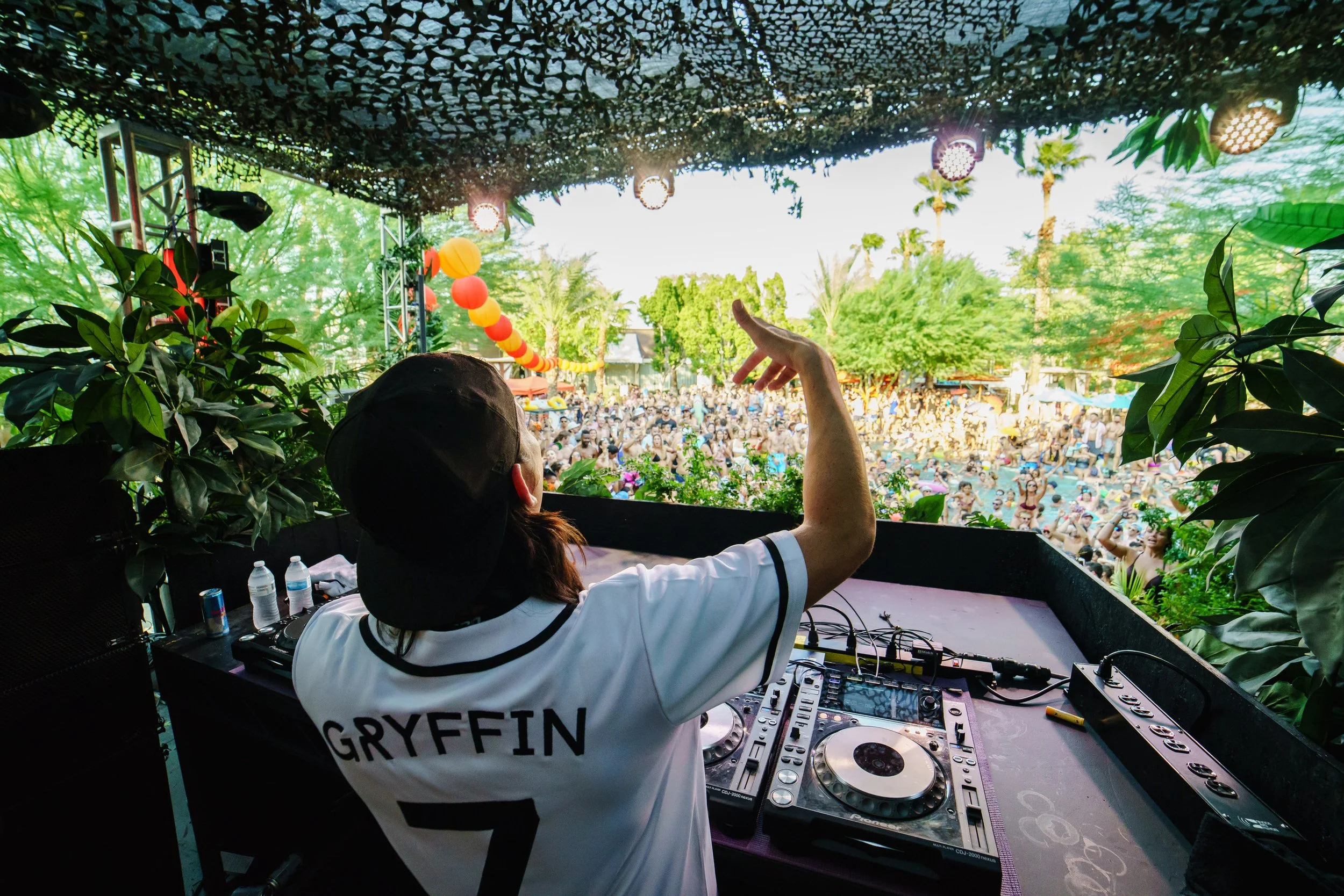 A DJ with the name Gryffin performing at an outdoor music festival, viewed from behind, with a large crowd and lush green trees in the background.
