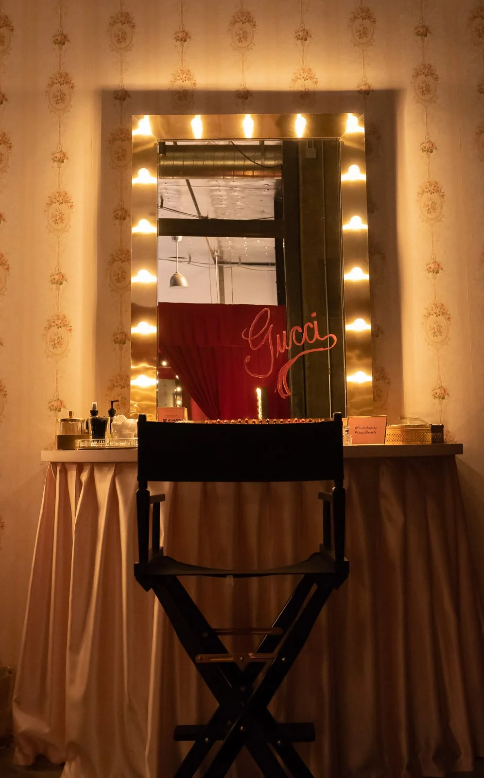 A vanity setup with a black director's chair, a large mirror surrounded by warm lights, and a table with skincare products, soap, and a sign in front of a floral wallpapered wall, with a red curtain reflected in the mirror and the name 'Guccì' writte