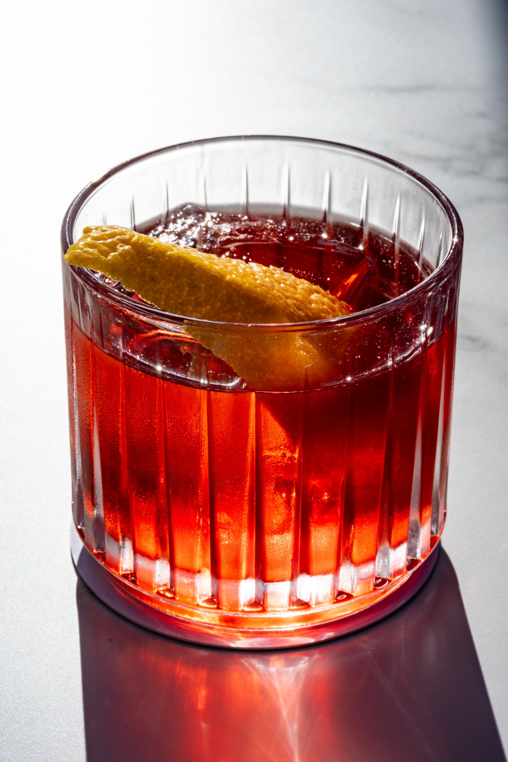 A tall glass with vertical ridges filled with a red beverage, garnished with a lemon peel twist.