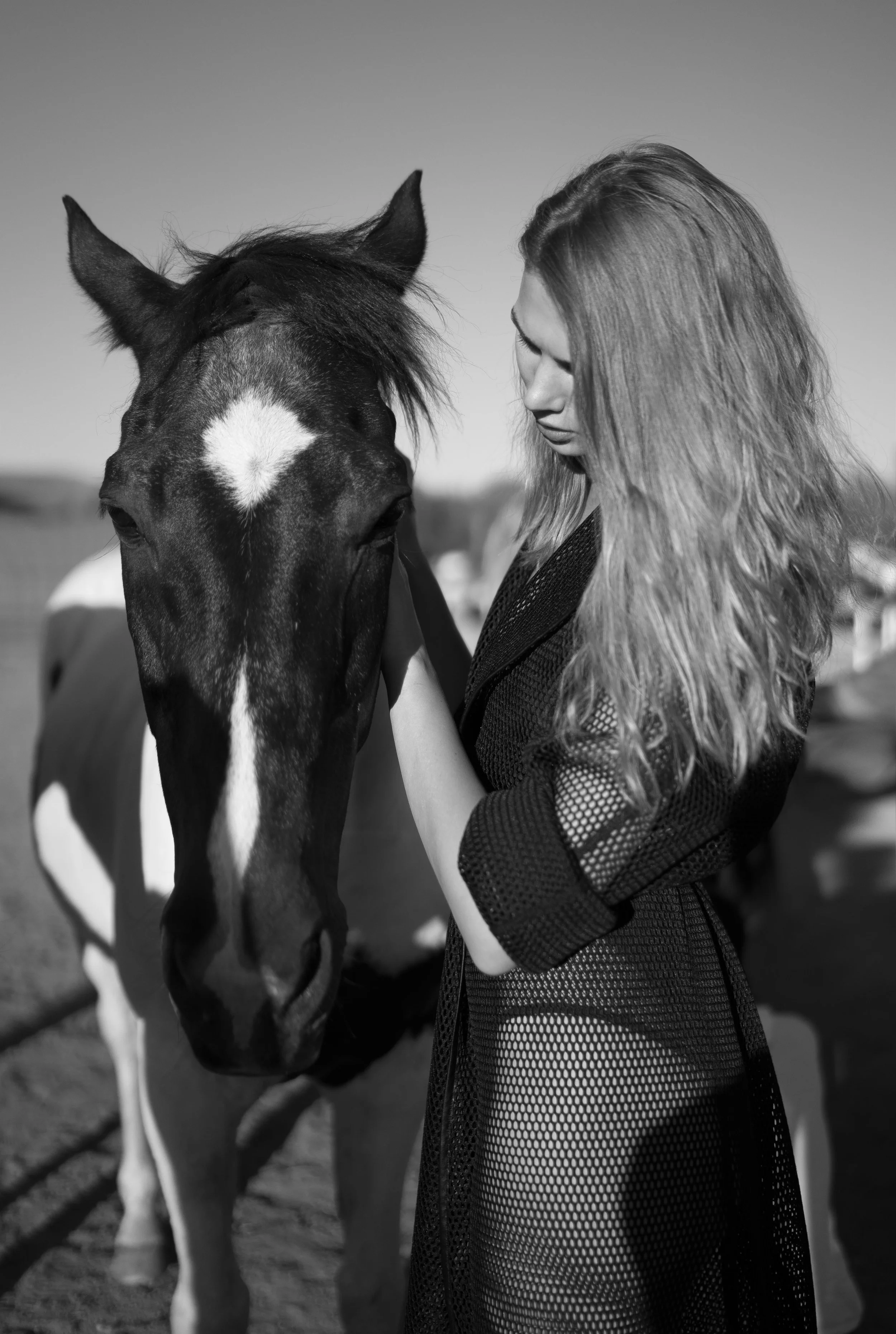 A woman with long wavy hair standing close to a horse. She is gently touching the horse's face, and both are facing each other.