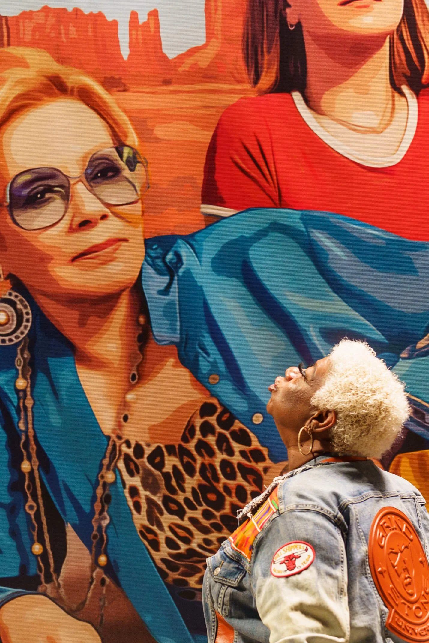 A woman with blonde hair and earrings is looking up and smiling, dressed in a denim jacket with patches and wearing a scarf. She is standing in front of a colorful mural of two women in a desert landscape. The mural features a woman with sunglasses a