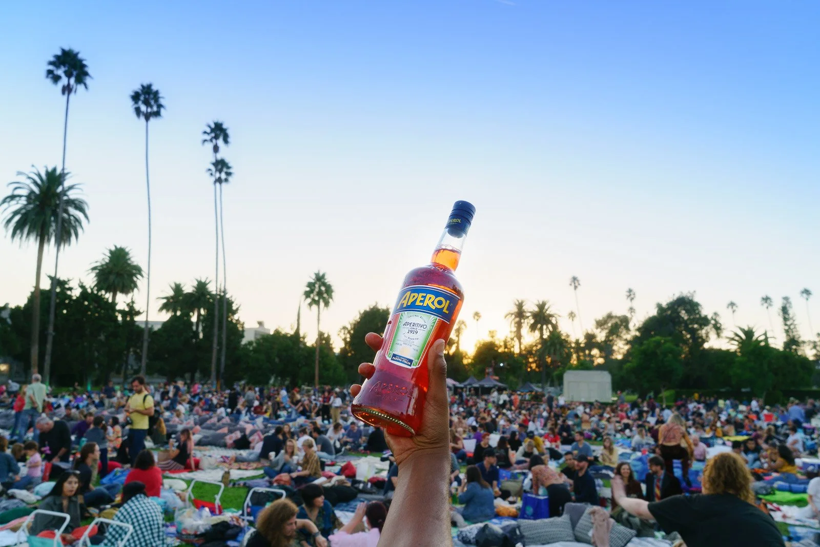 Hand holding a bottle of Aperol in front of a large crowd at an outdoor event during sunset, with palm trees and a clear sky in the background.