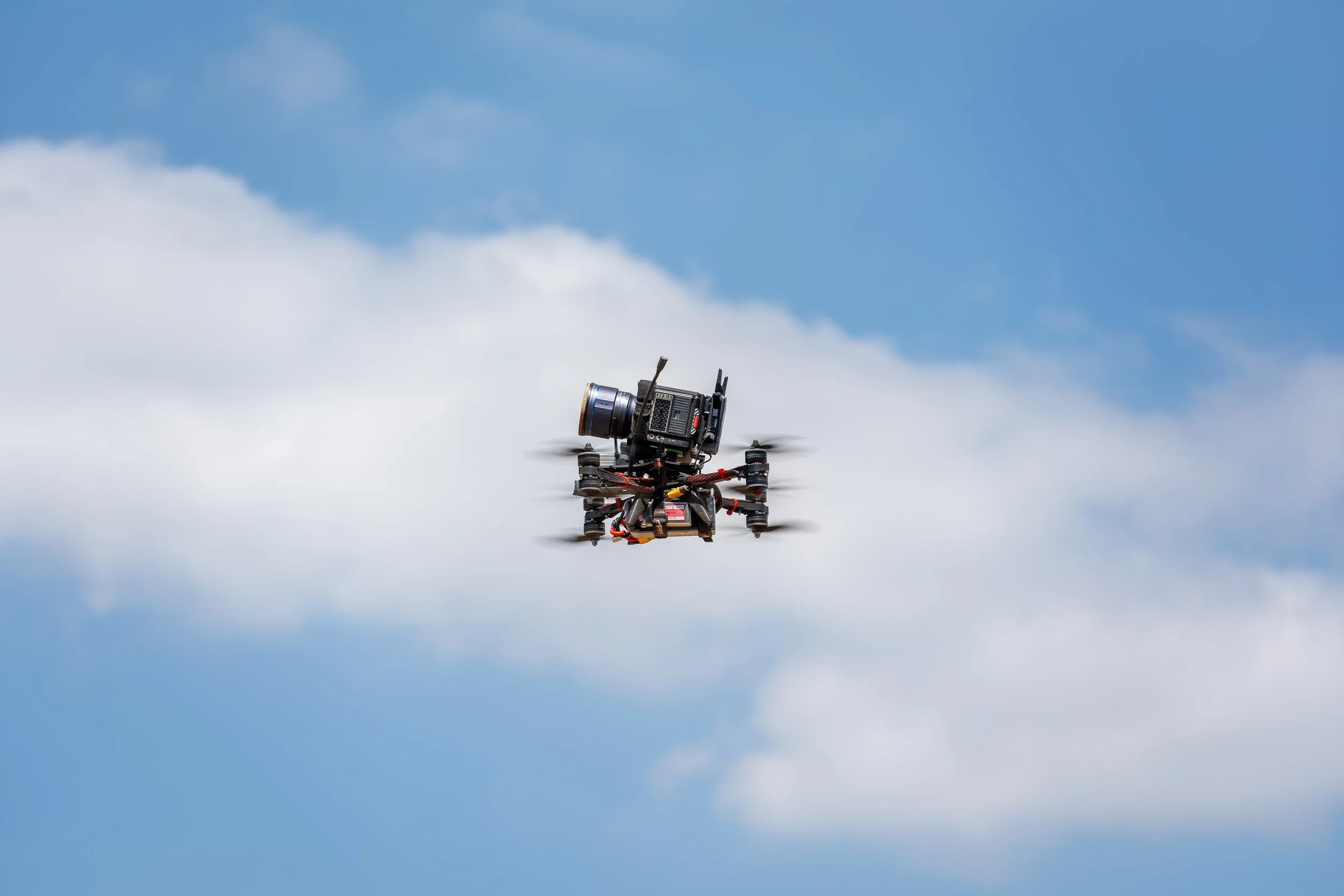 A drone flying in the sky with a camera attached to it, amid blue skies and white clouds.