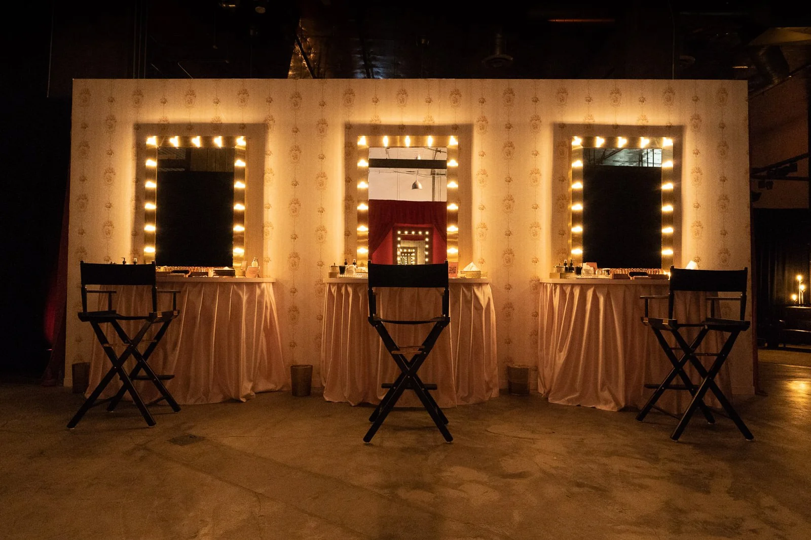 A beauty or makeup station with three mirrors surrounded by lights, each with a makeup chair in front. The background features a repeating floral wallpaper pattern, and the setup is on a wooden floor with soft ambient lighting.