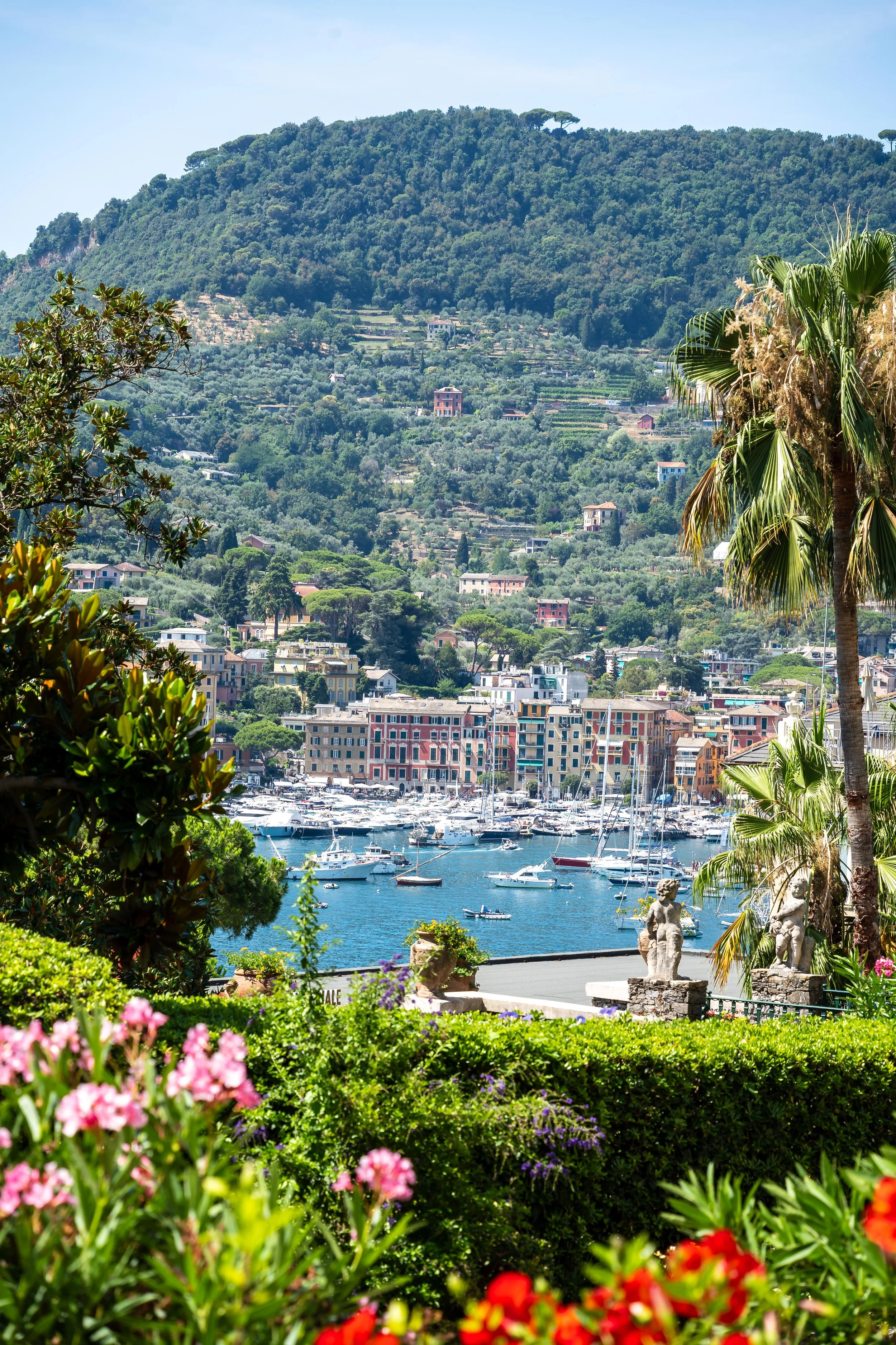 A marina with sailboats and yachts docked in calm blue water, colorful buildings on the waterfront, lush green hillside with houses and trees, and decorative statues among vibrant flowers in the foreground.