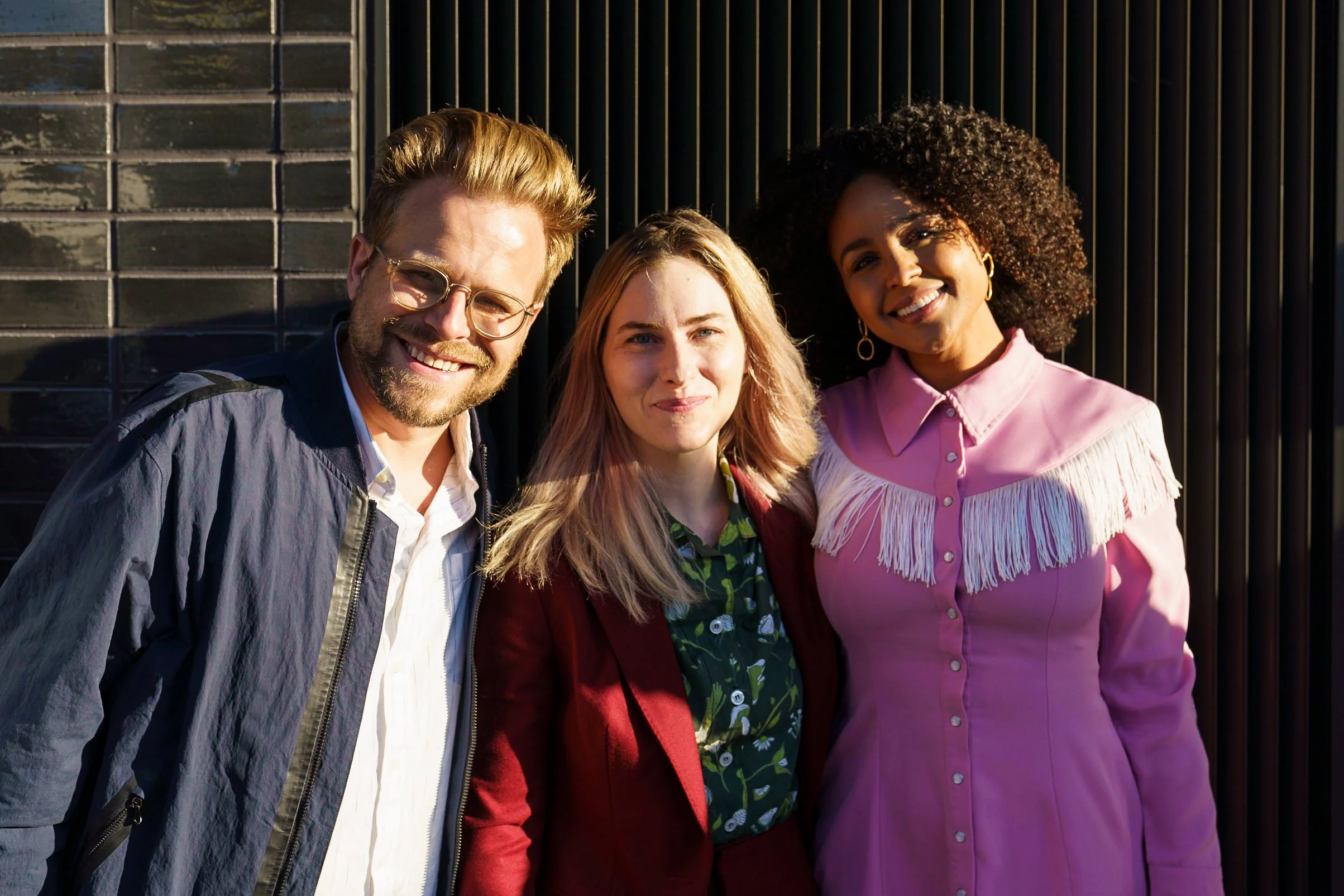 A group of three diverse young adults smiling and standing close together against a black fence with sunlight on their faces at HBO Hacks event.