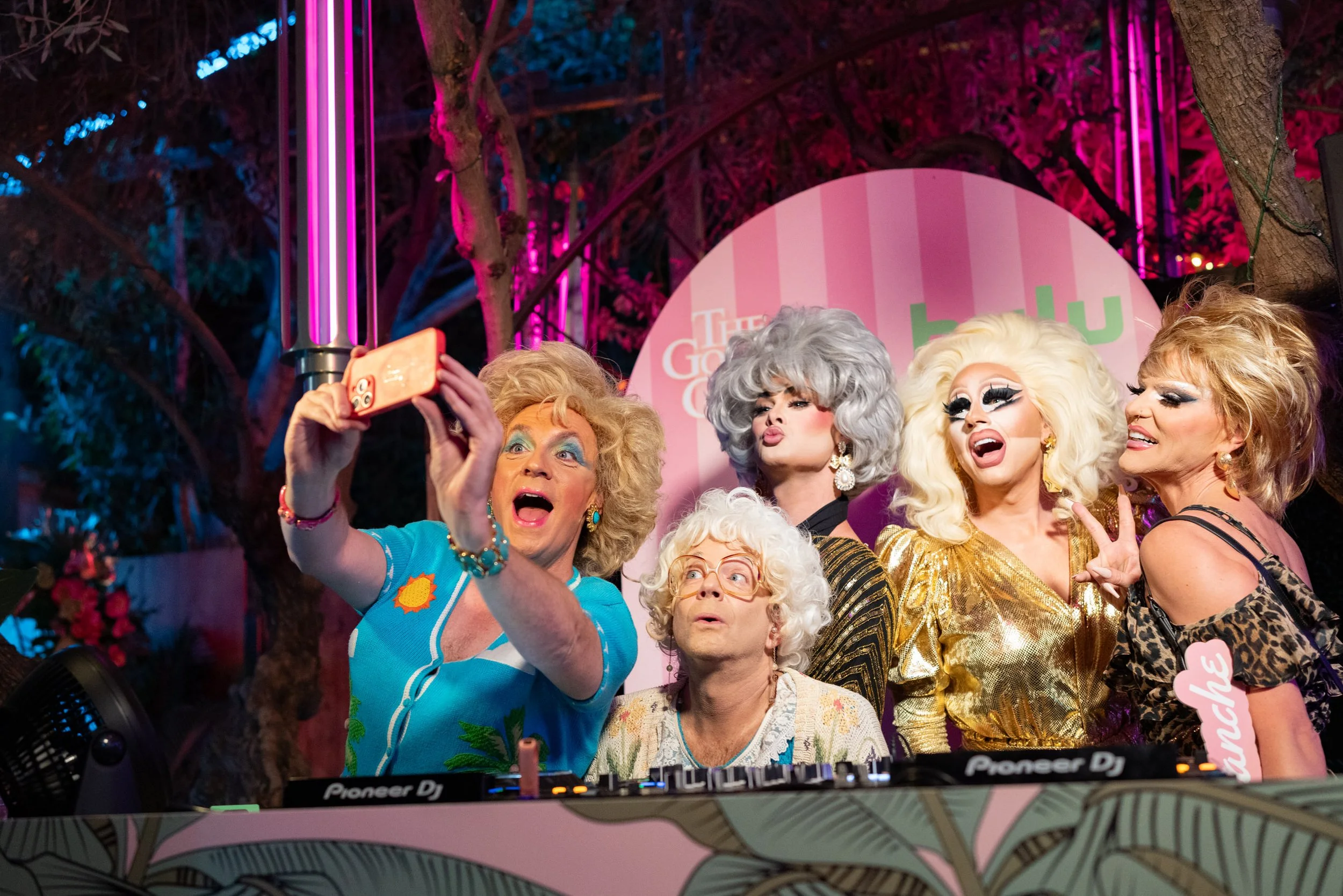 A group of drag queens taking a selfie at an event with a pink backdrop and neon lights, surrounded by trees for HULU's Golden Girls 40th Anniversary.