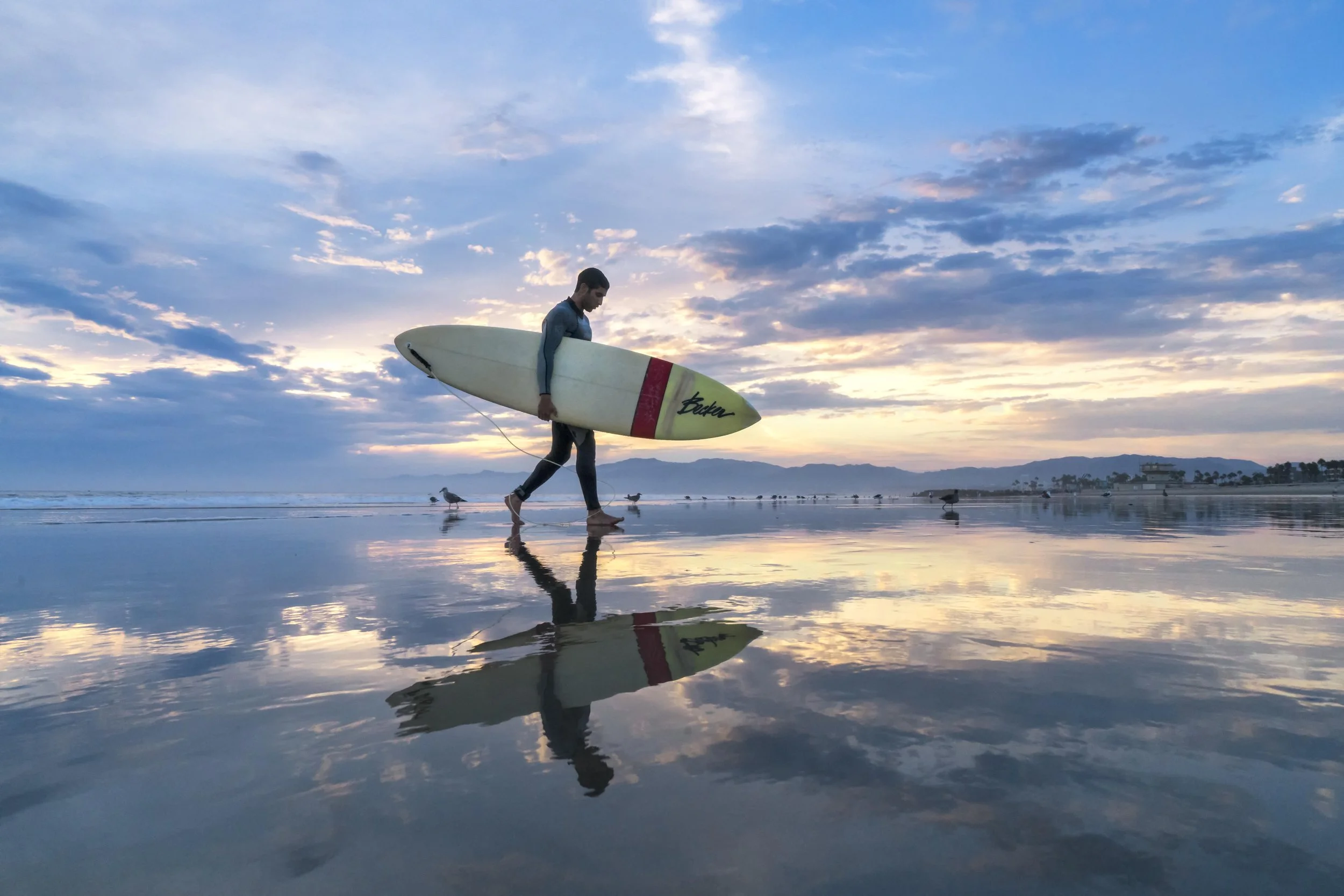 A man walking on the beach carrying a surfboard at sunset, with clouds in the sky and reflections on the wet sand.