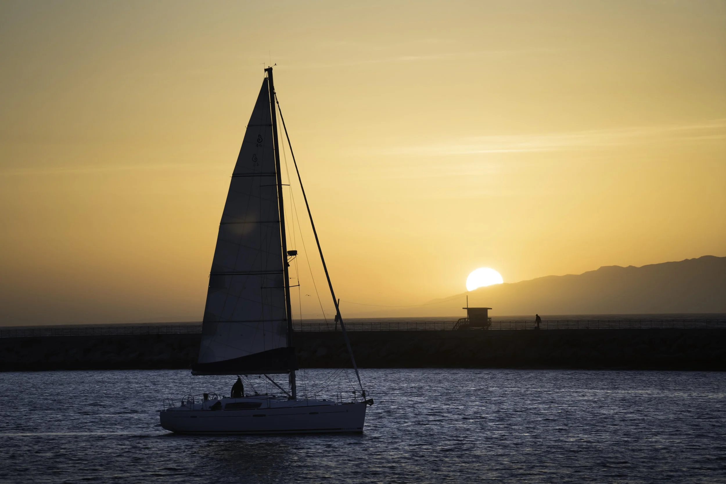 Sailboat on the water during sunset with a person walking on a pier in the background in Marina Del Rey, Los Angeles, California.