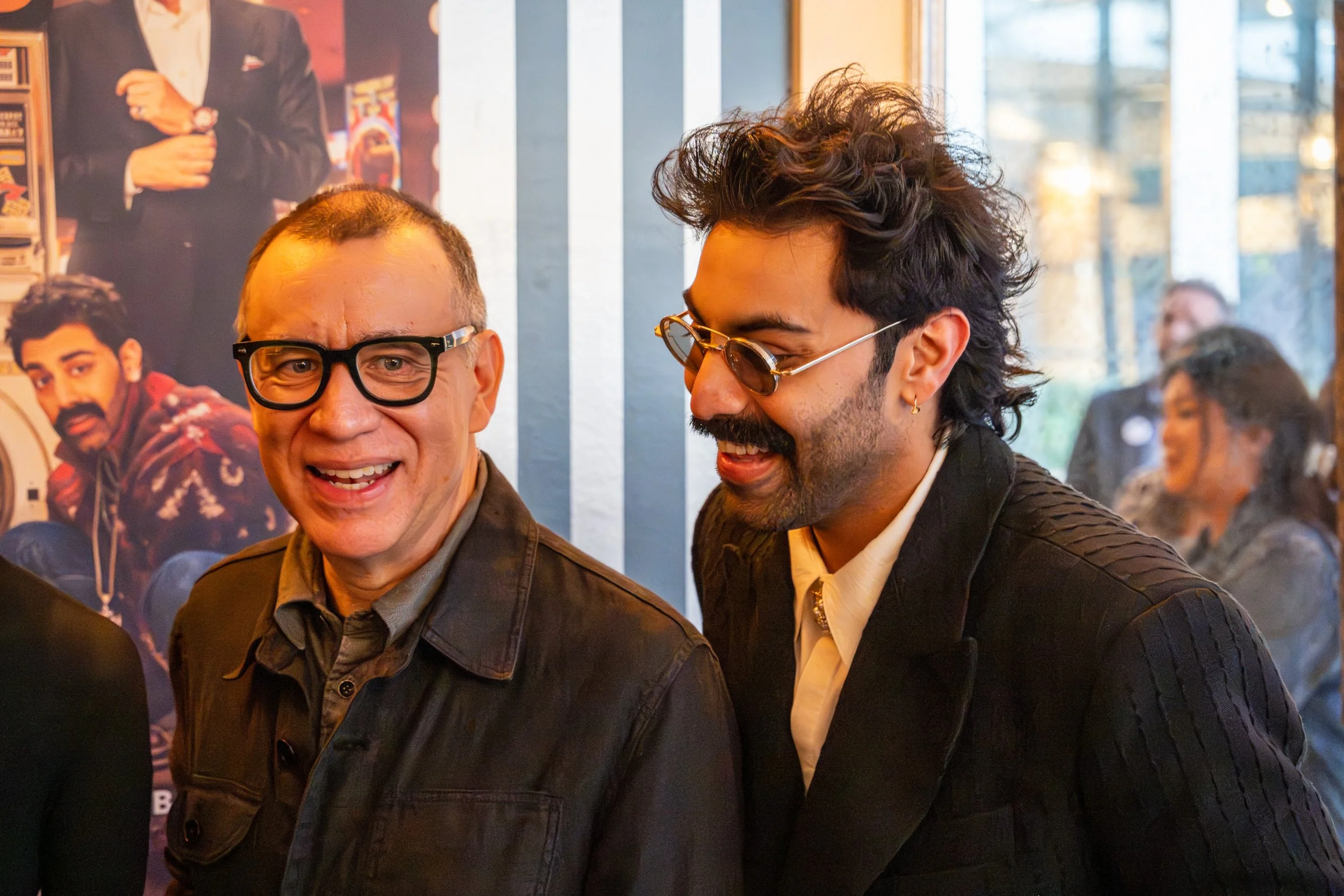 Actors Fred Armisen and Saagar Shaikh are smiling and appear to be talking, in an indoor setting with posters on the wall behind them at HULU's Season 2 party for Deli Boys TV show.