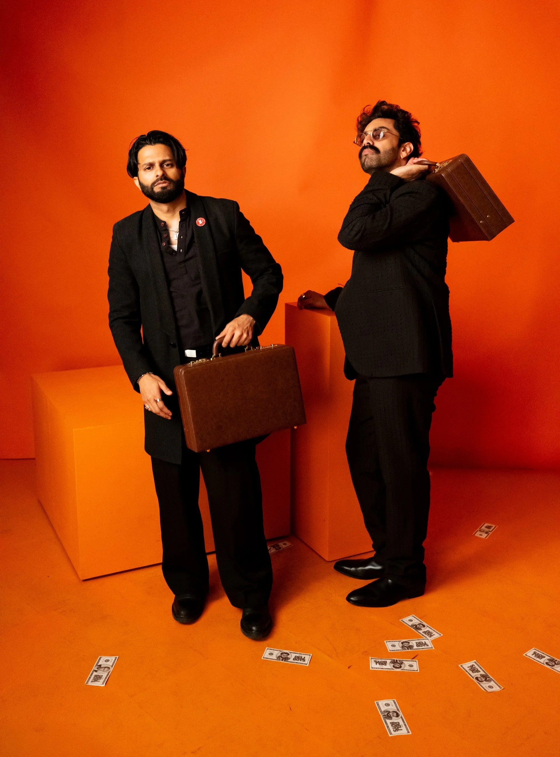 Two men in black suits stand in front of an orange background, holding brown briefcases. One man has a serious expression, and the other is facing sideways. Fake US dollar bills are scattered on the orange floor.