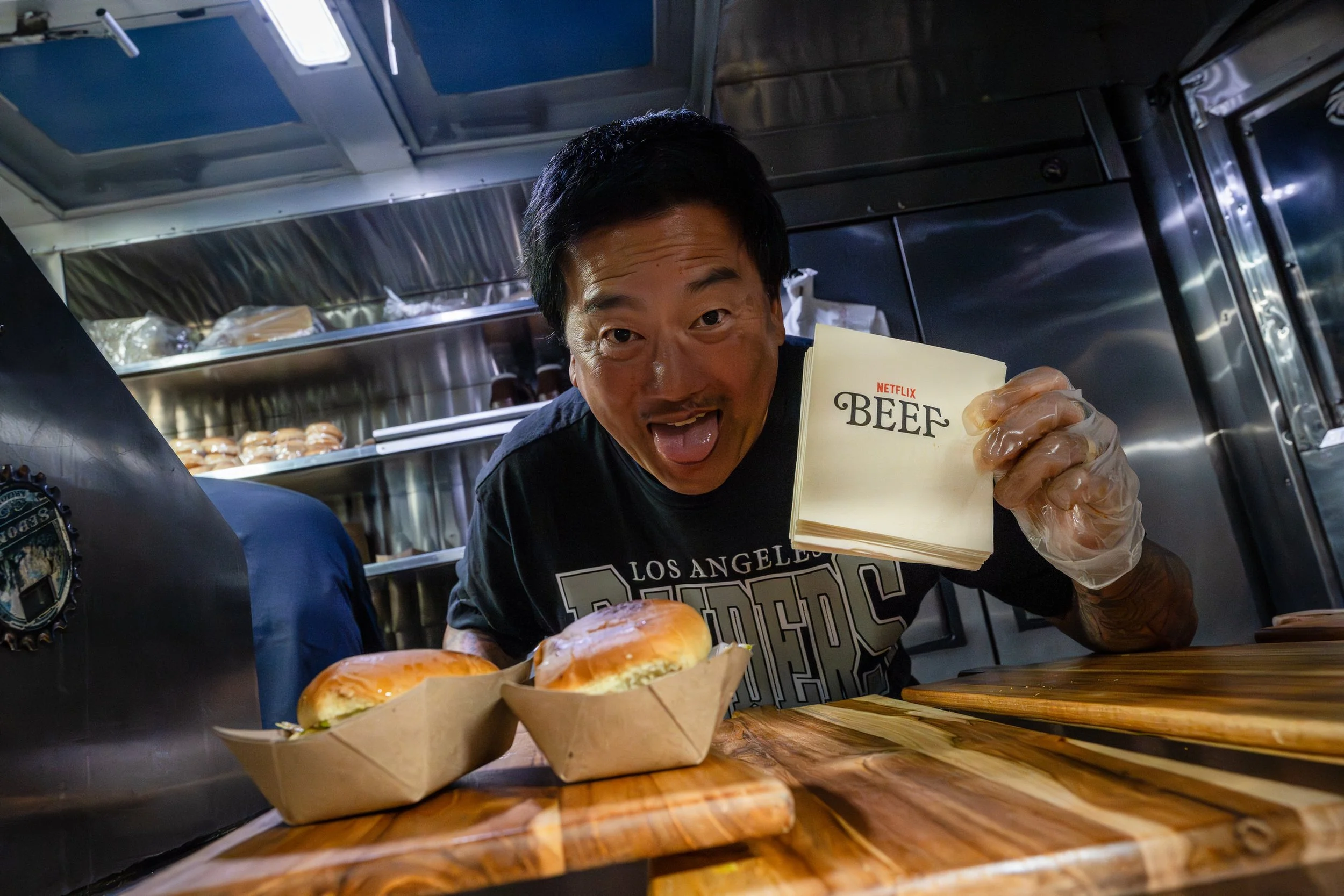 A man holding a napkin with the word 'BEEF' printed on it, smiling at the camera in a food truck. The food truck interior is metallic with shelves of food and pastries at NETFLIX's BEEF premiere event.