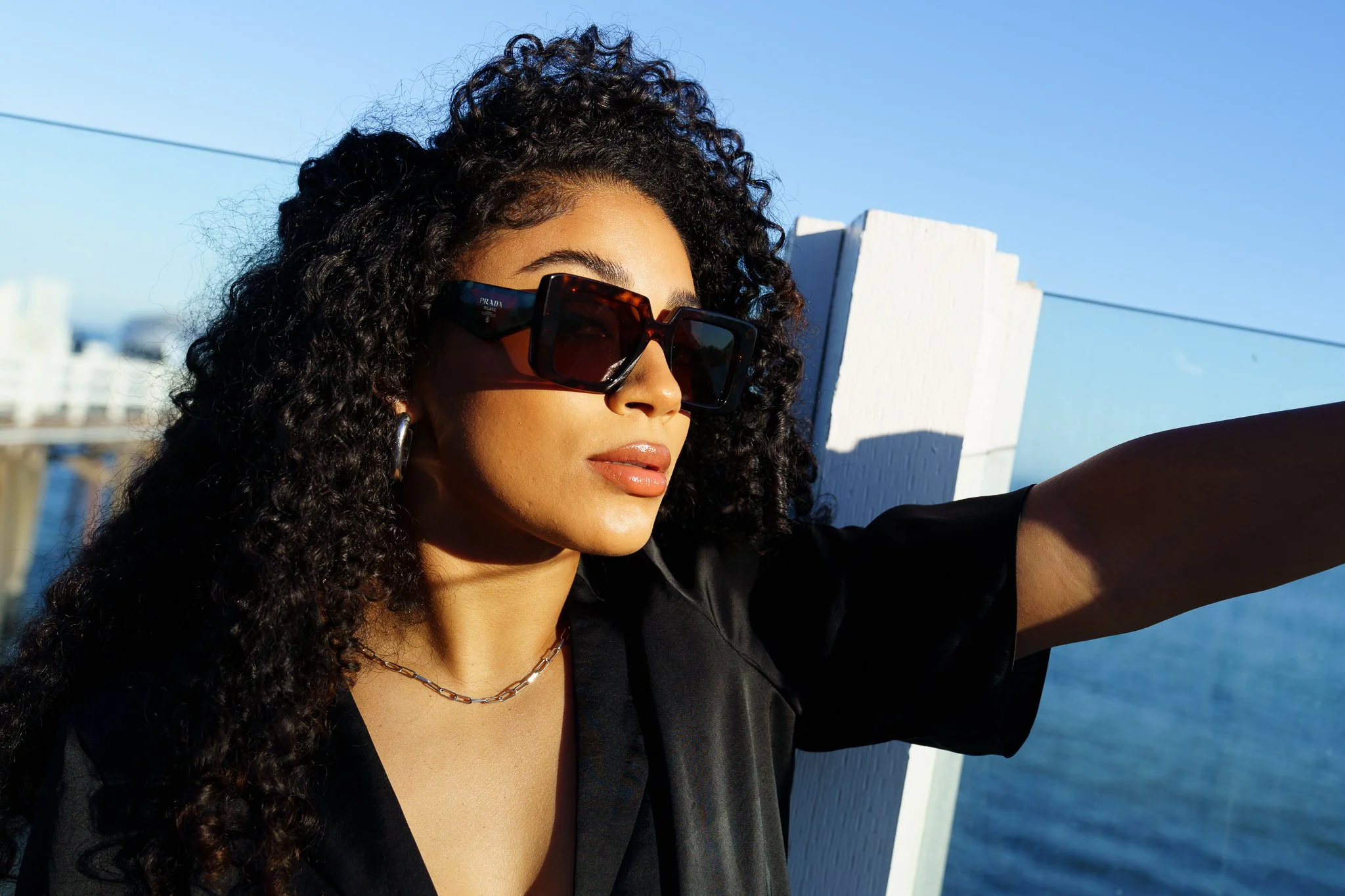 A woman with curly black hair and large Prada sunglasses taking a selfie outdoors near a body of water and white fence, with a clear blue sky in the background. Sunglass Hut event at Malibu Pier.