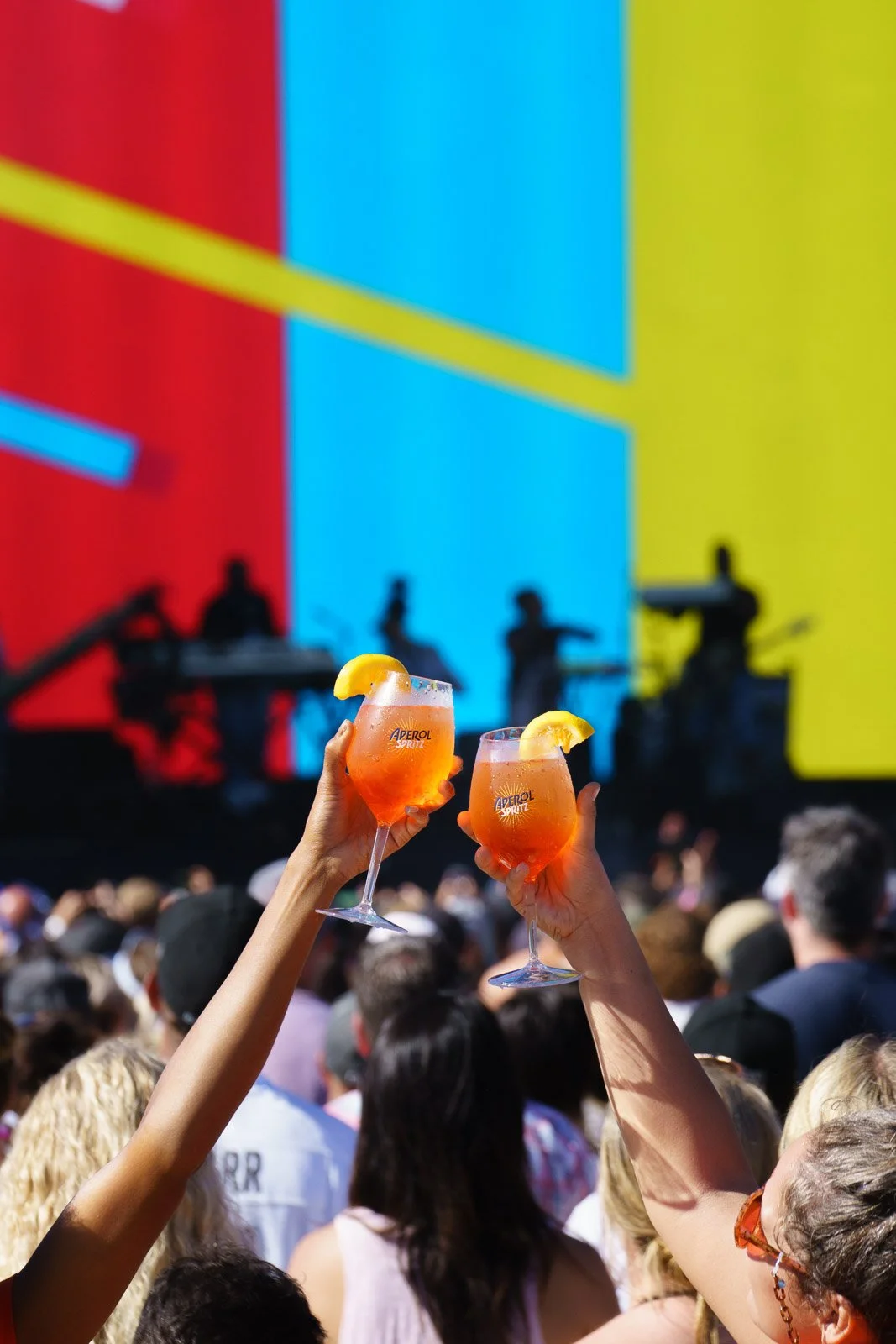 Two hands holding glasses of orange Aperol Spritz with lemon slices, raised in a toast at an outdoor concert with a large colorful stage and a crowd of people.
