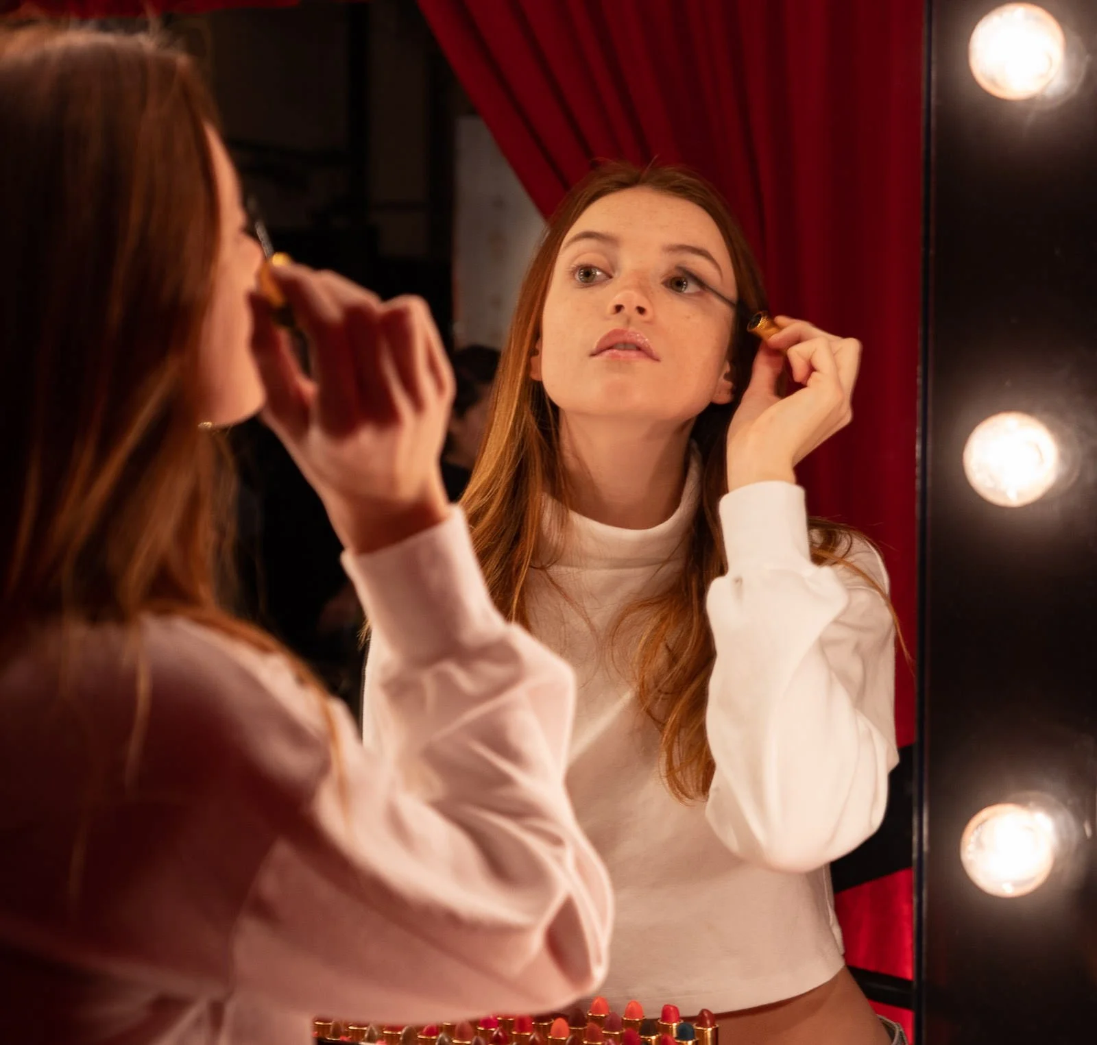 A young woman with red hair applying makeup in front of a mirror with lightbulbs. Red curtains are in the background.