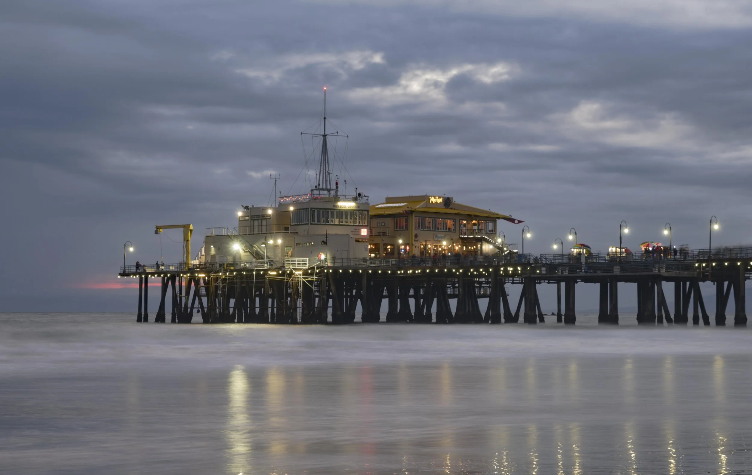 Santa Monica Pier with a building and outdoor tables, illuminated with string lights, extends into the ocean under a cloudy sky during sunset or dusk.