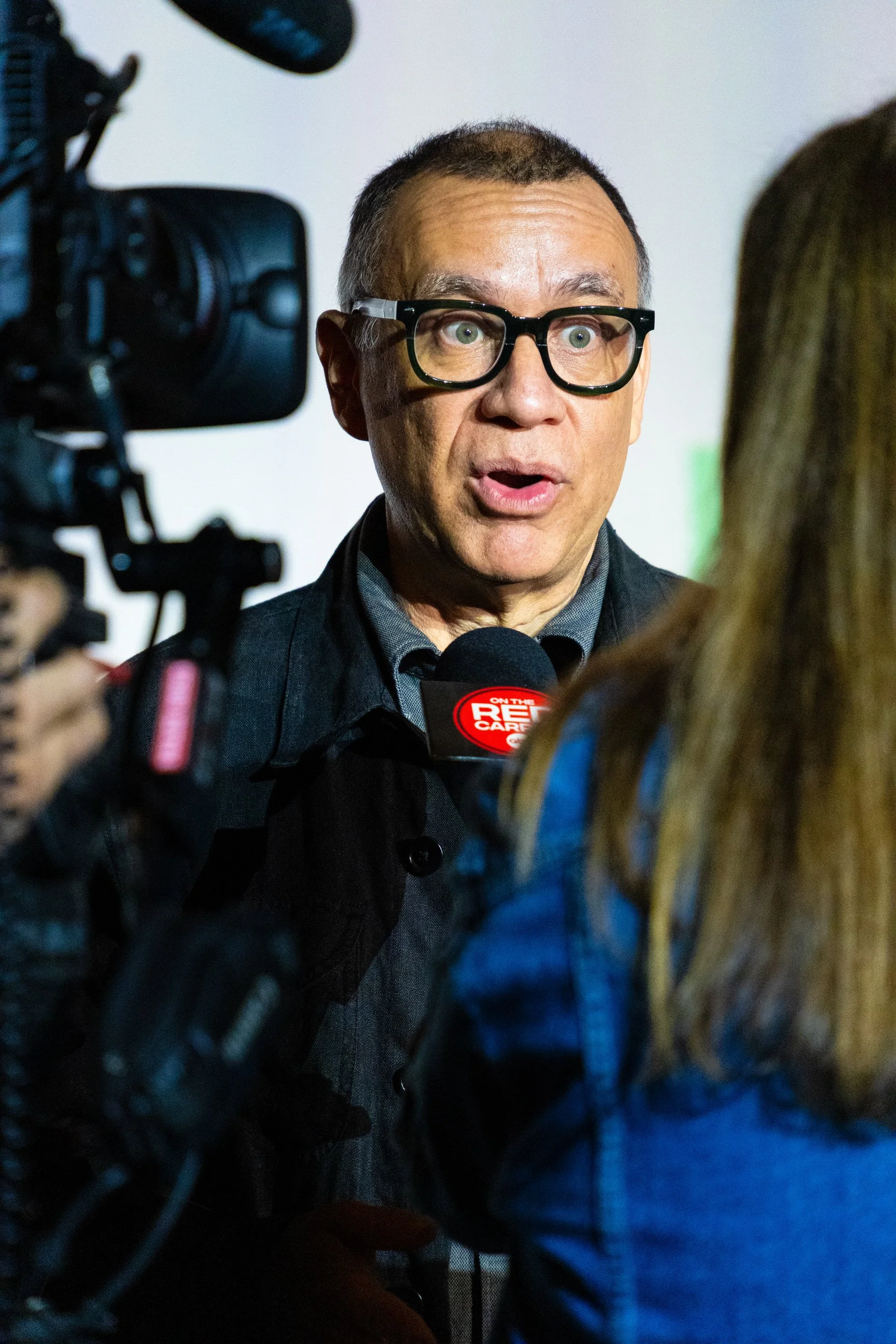 Actor Fred Armisen with short hair, glasses, and a dark jacket being interviewed by a woman with long hair. A camera and microphone are visible, suggesting a news interview setting at the premiere for HULU's Deli Boys Season 2.