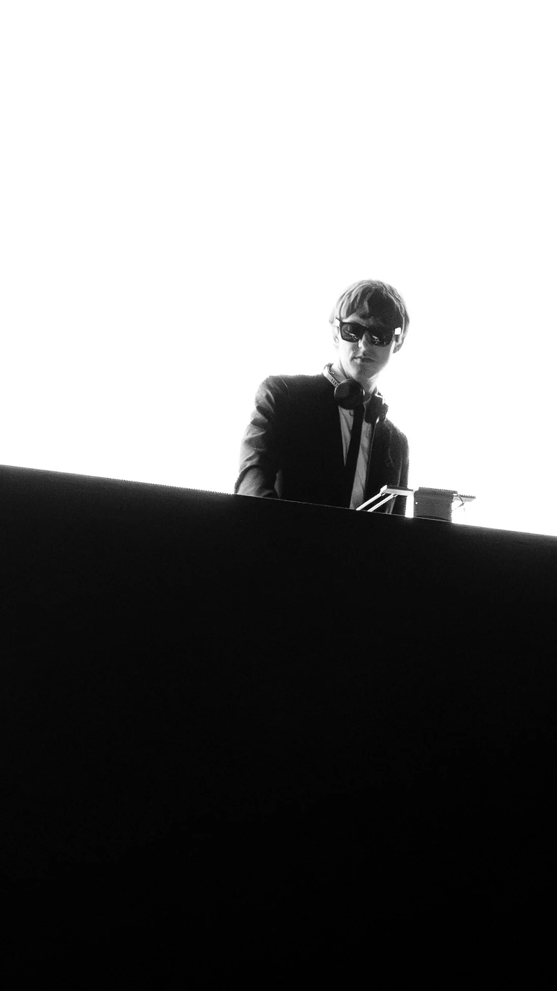 Producer and DJ The Dare wearing sunglasses, headphones around neck, and a blazer standing behind a DJ booth, with a bright white background at NETFLIX's Beef Season 2 premiere party.
