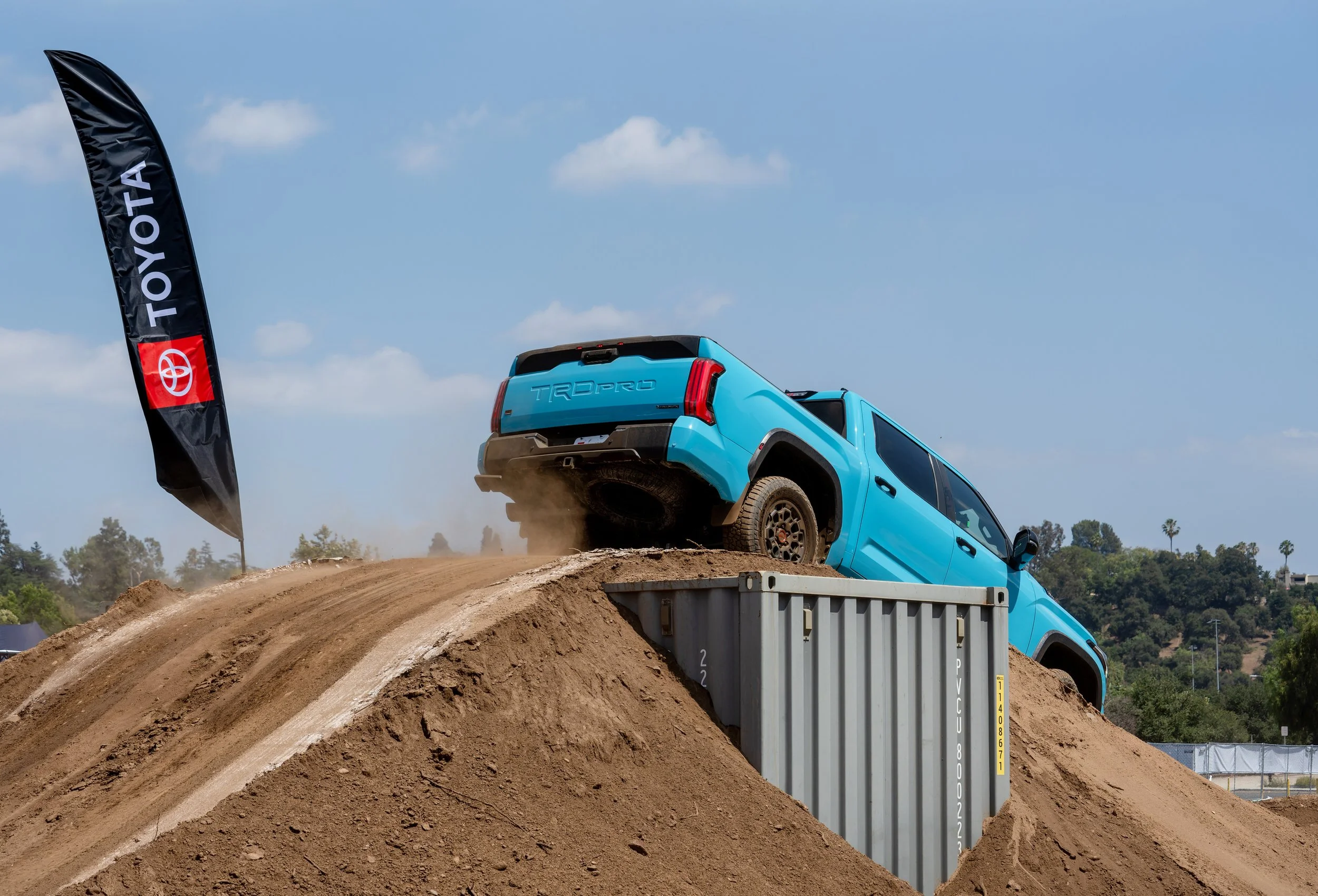 A blue Toyota Tundra TRD Pro truck climbing a dirt hill during an off-road event, with a black Toyota flag on the left side of the scene and a clear sky with some clouds in the background.