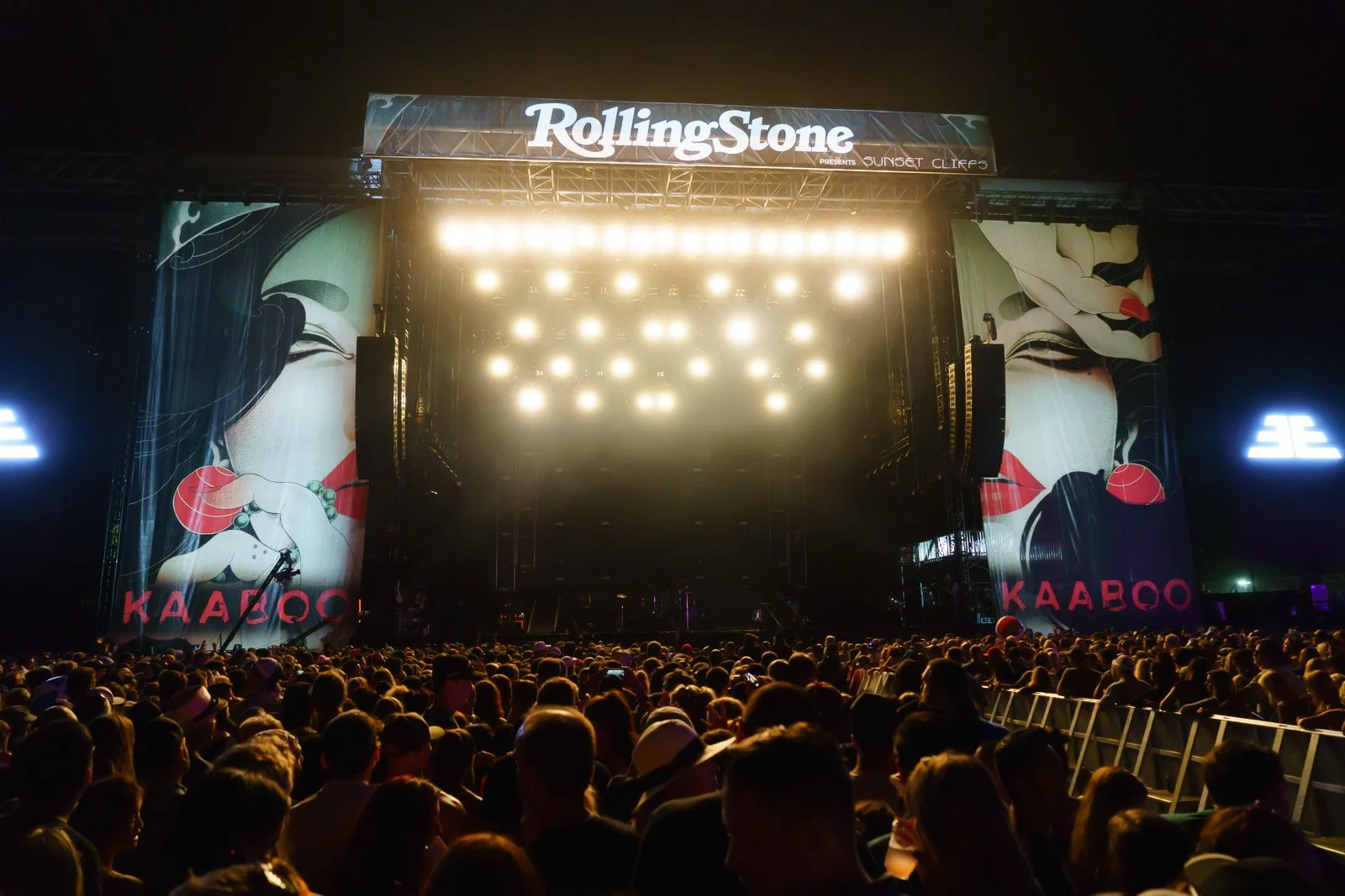 An outdoor concert stage with bright lights and a large crowd at night. The stage has a Rolling Stone sign at the top and artistic banners on the sides featuring stylized images of women with red lips and cherry accessories, with text in a non-Latin 