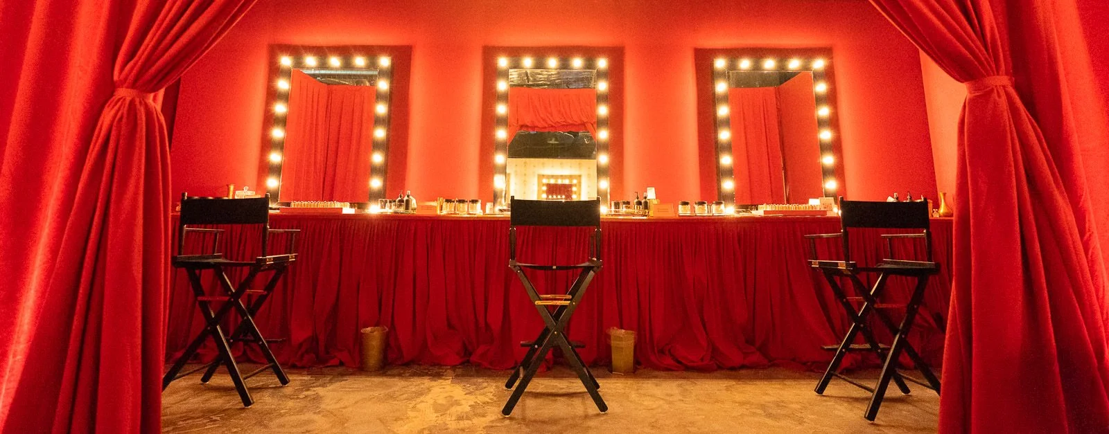 A makeup or dressing room for Gucci Beauty surrounded by bright lights. The room is decorated with red curtains and a red tablecloth. There are three black chairs in front of the mirrors and various makeup or beauty products on the table.