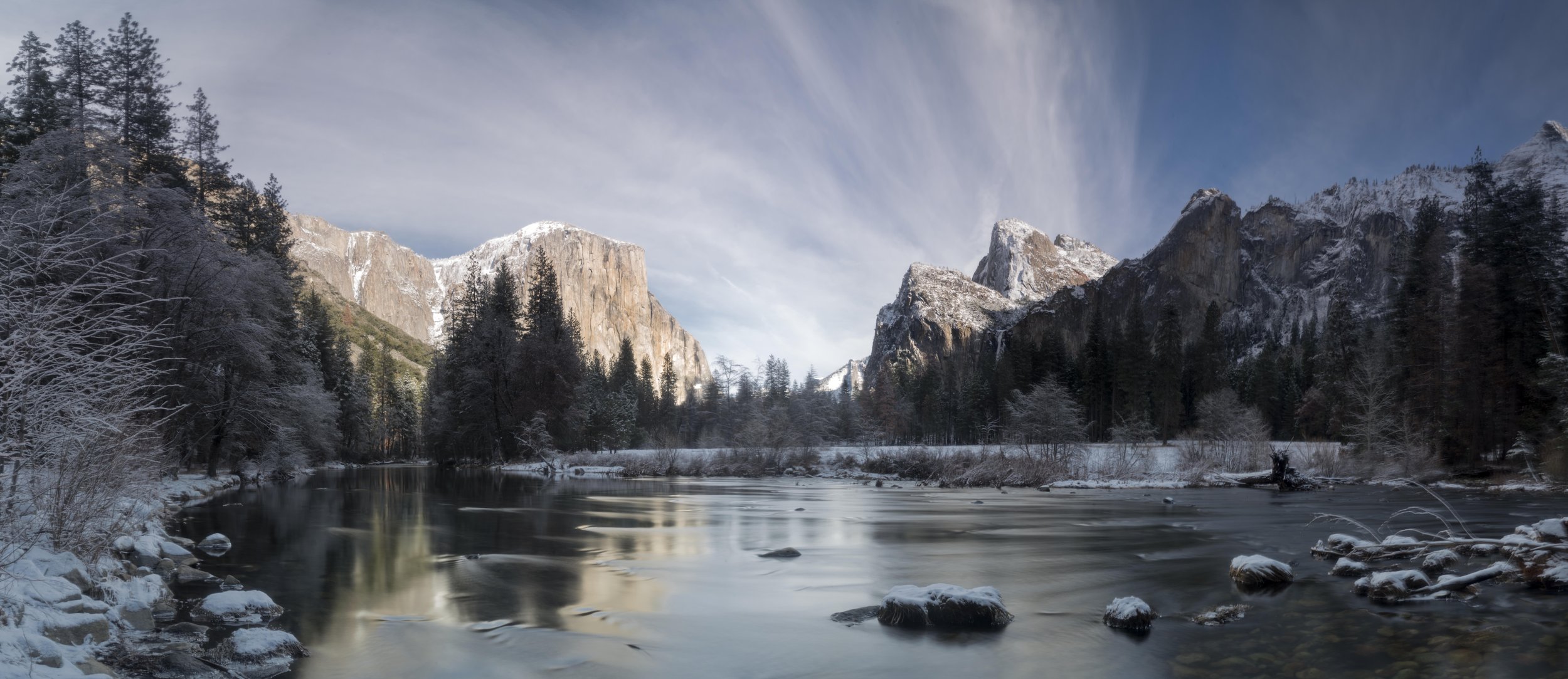 Snow-covered trees and mountains surrounding a calm river in a winter landscape.