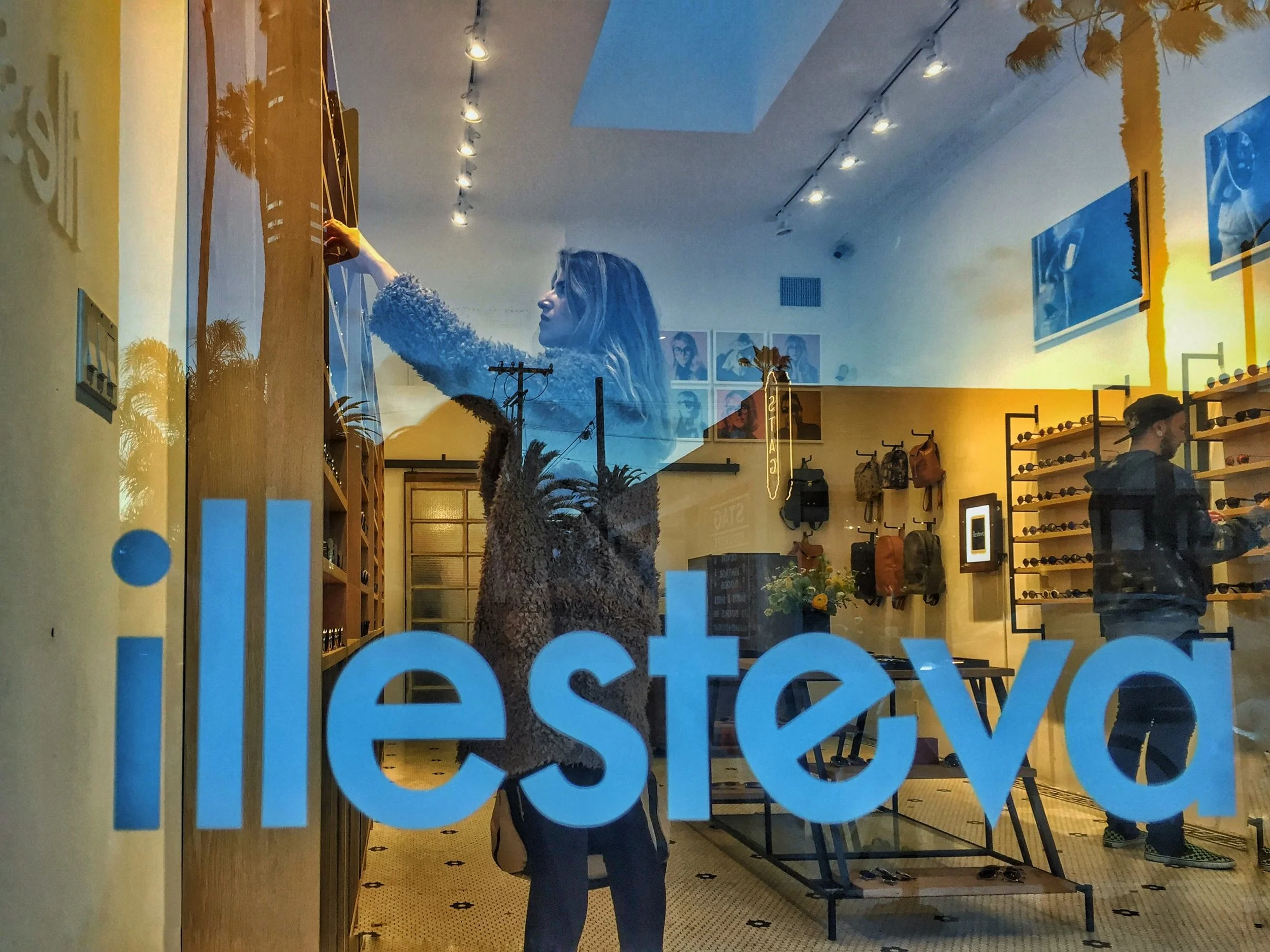 A woman with long hair and a fuzzy sweater is reaching up to a display on a shelf inside a store, reflected in the store window. The store has shelves with bags and sunglasses, and framed pictures or artwork on the wall. The photo is taken from outsi