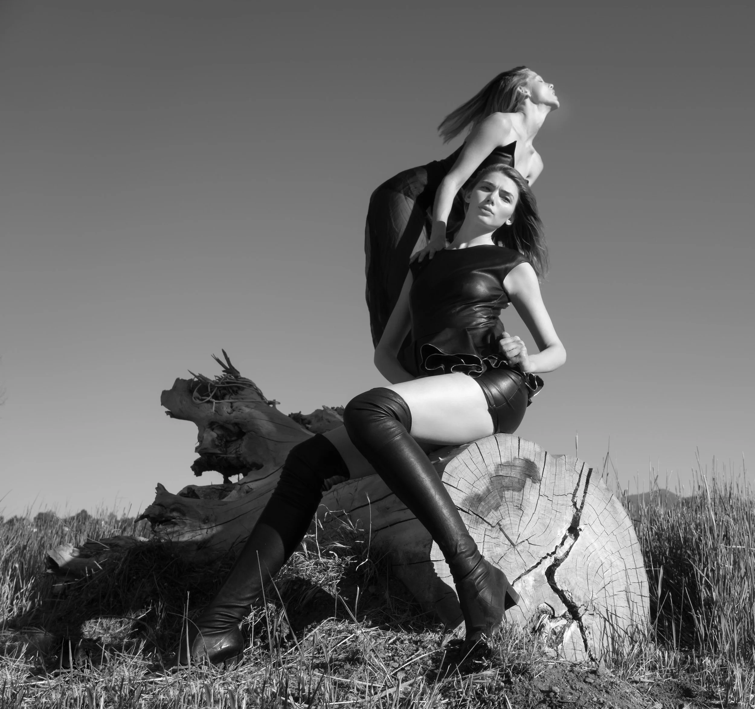 Black and white photo of two women outdoors on a fallen log, with one sitting and one kneeling, both looking up and to the right, under a clear sky.