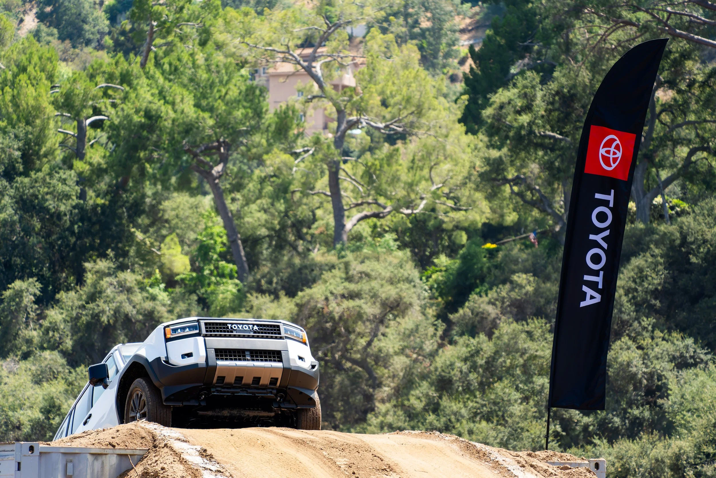 A white Toyota off-road vehicle is positioned on top of a dirt ramp or mound, with a backdrop of a forested hillside and a building in the distance. A black vertical banner with a red and white Toyota logo is visible on the right side of the image. T