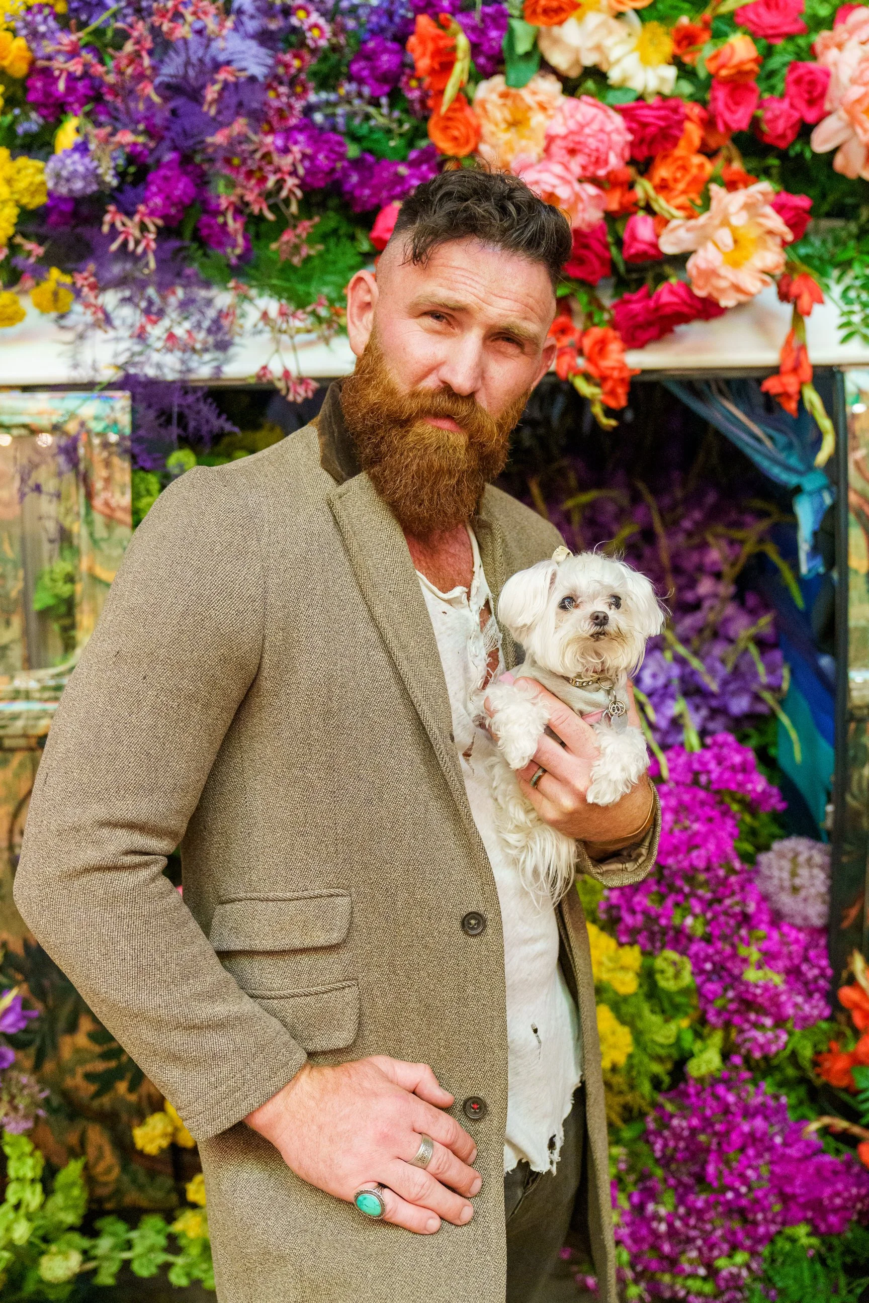 Actor Devan Chandler Long with a reddish beard and short hair wearing a beige blazer and white shirt, holding a small white dog, standing in front of a colorful flower display for CBS's Ghosts TV show.