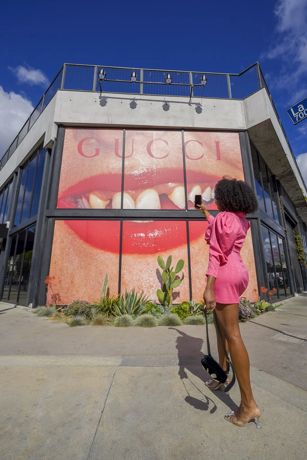 A woman in a pink dress taking a photo with her phone outside a building with a billboard of a close-up of a mouth with red lipstick, teeth, and the word "Gucci" at the top.  For Gucci Beauty.