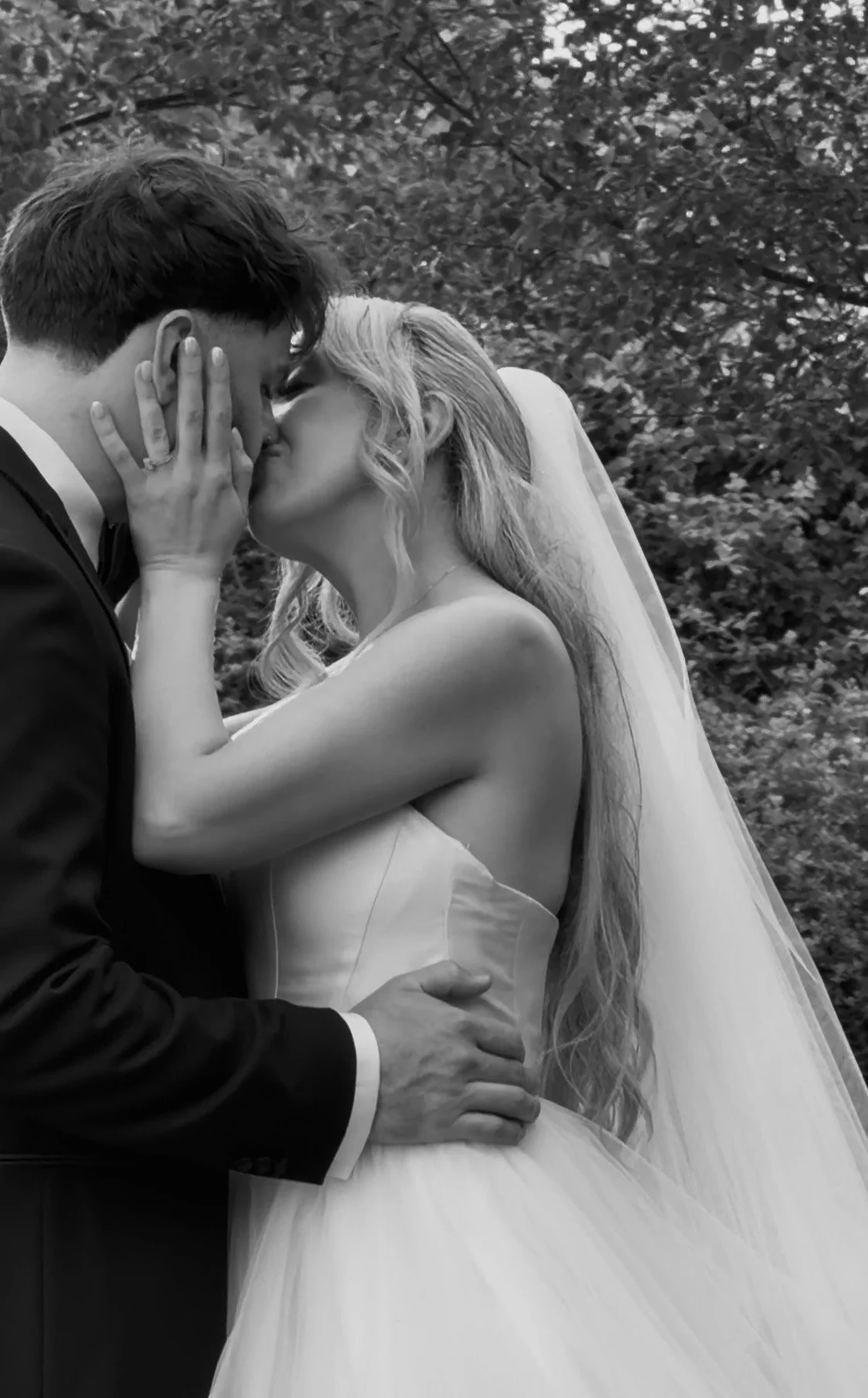A black-and-white photo of a bride and groom sharing a kiss outdoors. The groom wears a tuxedo, and the bride wears a strapless wedding gown with a veil. She has her hand on the side of his face, and he gently holds her waist.