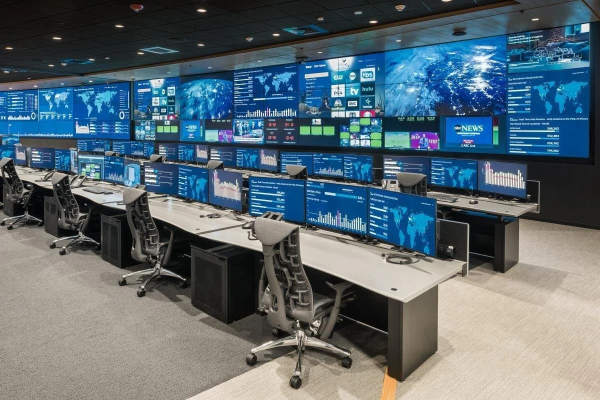 A large control room filled with multiple rows of workstations with computer monitors displaying graphs, maps, and data. The background wall features a series of large screens showing world maps, charts, and news info.