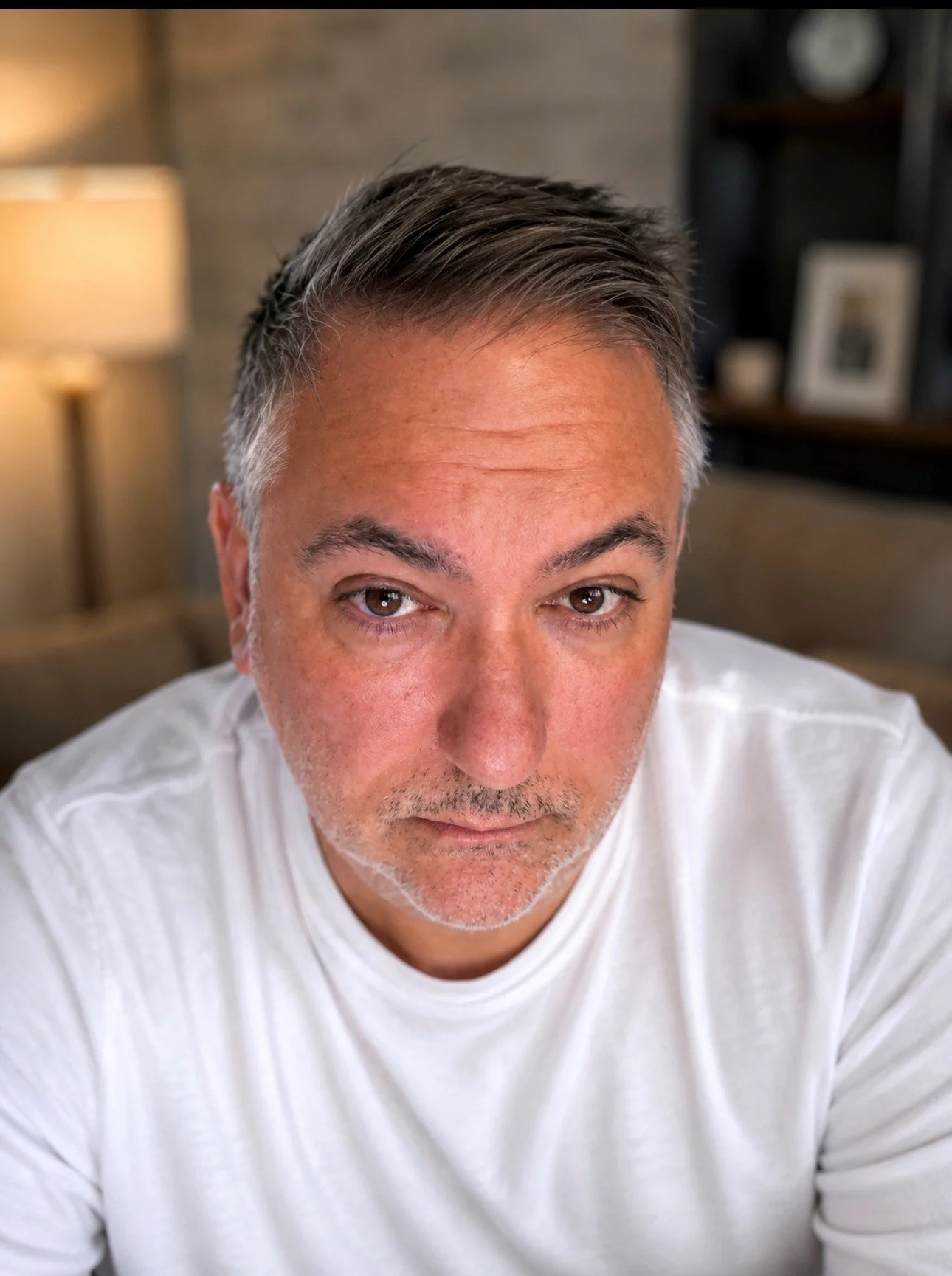 Close-up of a middle-aged man with short, gray hair, brown eyes, and light facial hair, wearing a white shirt, sitting indoors with a blurred background of a room with a lamp and framed photograph.