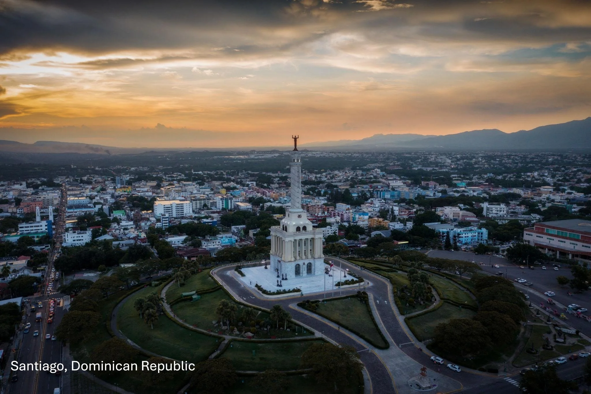 A cityscape of Santiago, Dominican Republic with a prominent white monument featuring a statue at the top, surrounded by a circular road and green landscape, under a colorful sky with clouds and sunset hues.