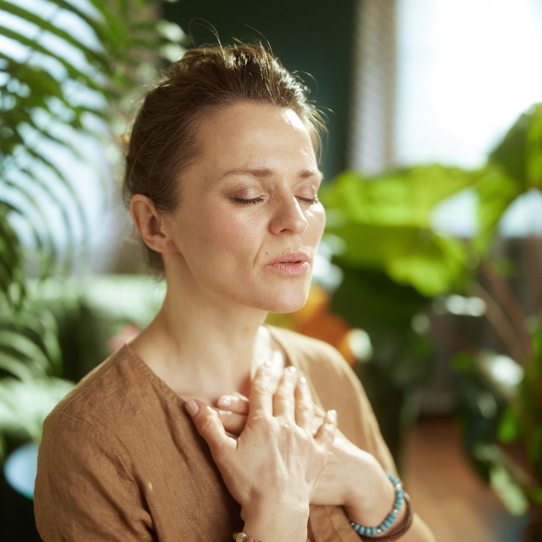 A woman with short brown hair and closed eyes, wearing a brown top, doing sukh tantra breathwork with hand on her chest, appearing to be calm or meditative.